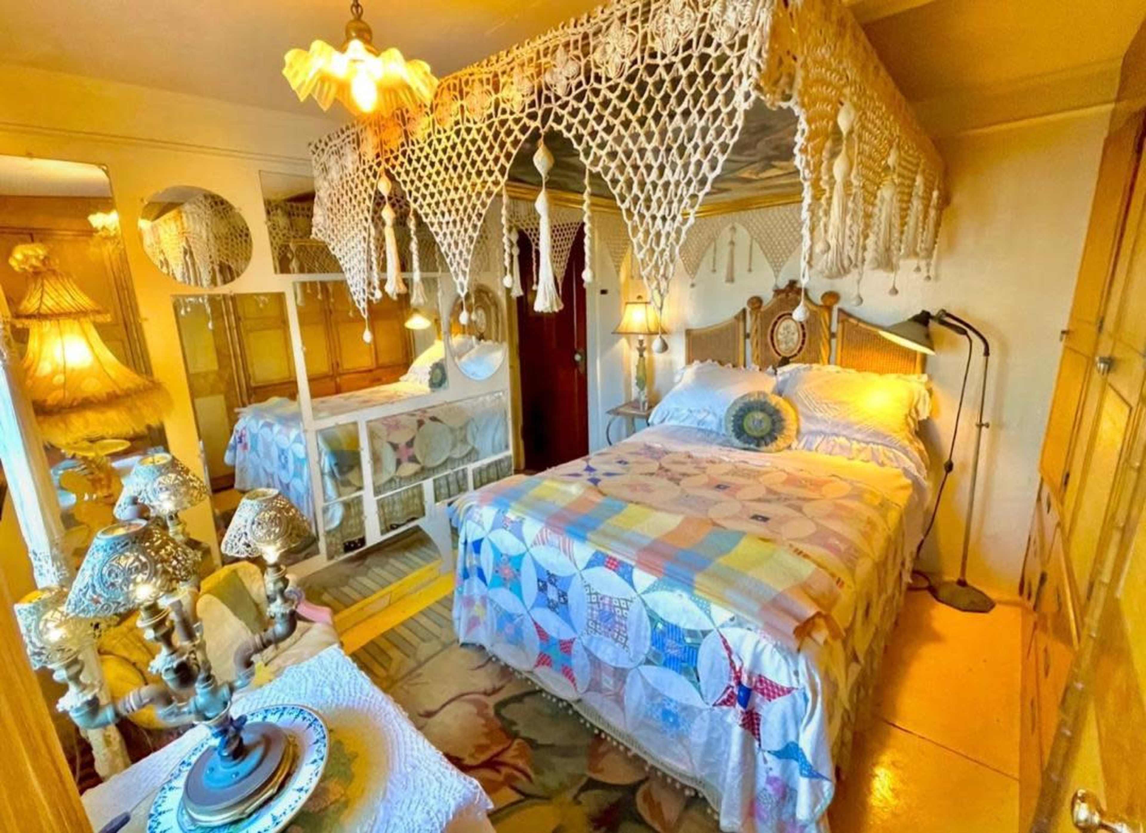 The image shows a cozy bedroom featuring a bed with a colorful patchwork quilt, a canopy made of lace, and various vintage lamps and decor items.