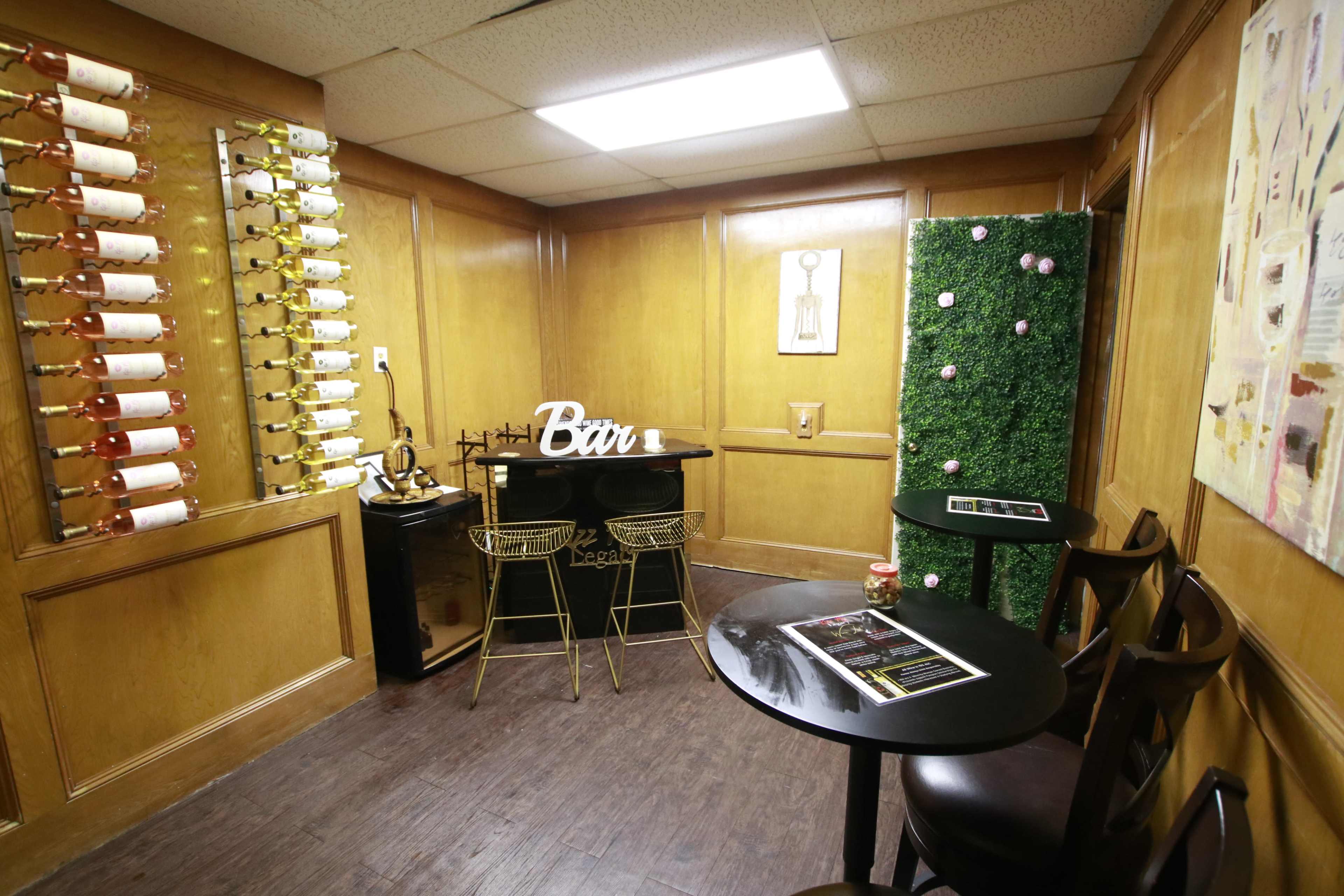 A small wooden-paneled room featuring wine racks on one wall, a black bar table with gold stools, and a decorative green wall.