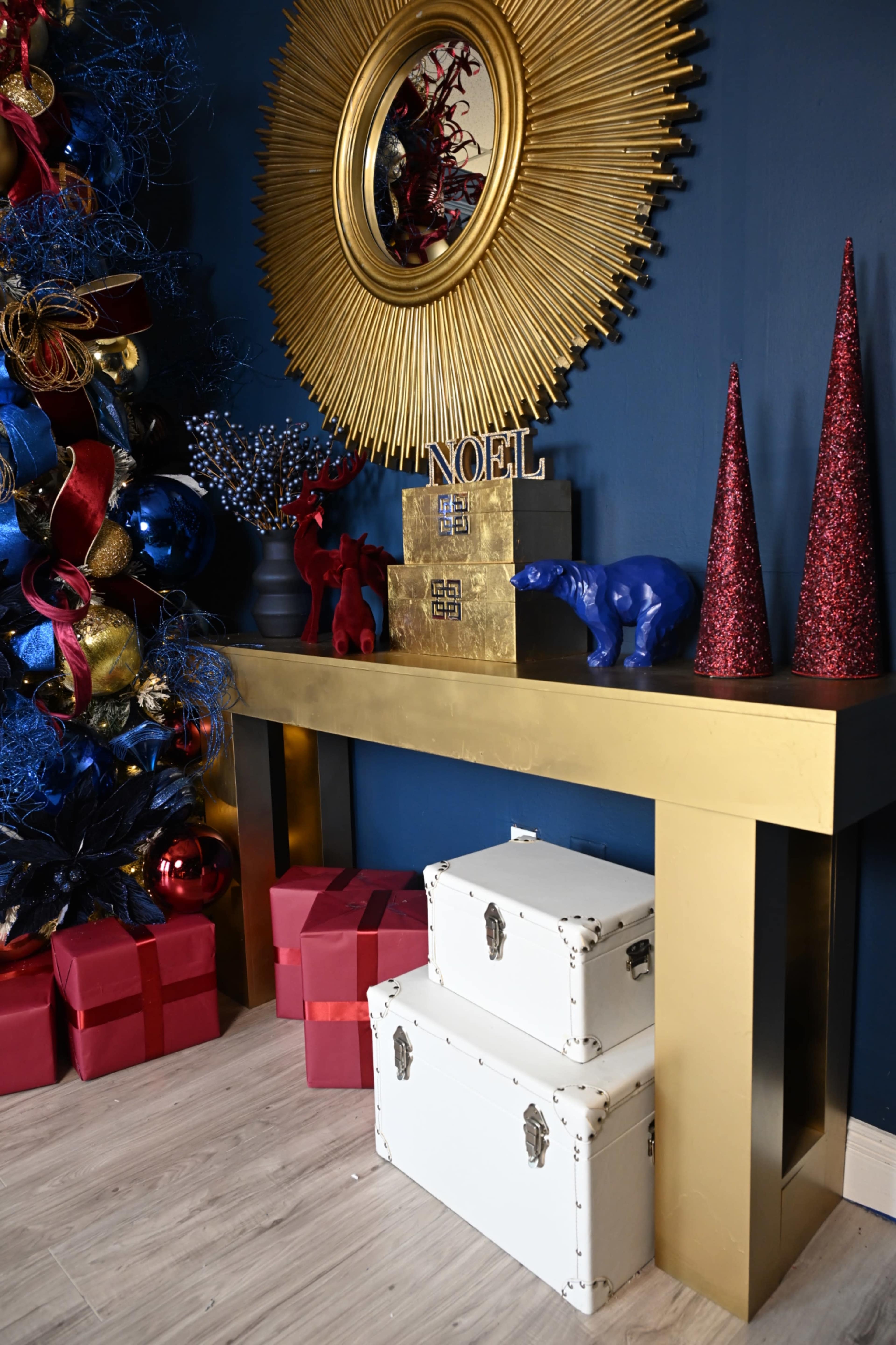 A gold console table is positioned against a dark blue wall, decorated with a sunburst mirror, colorful ornaments, and stacked gift boxes.