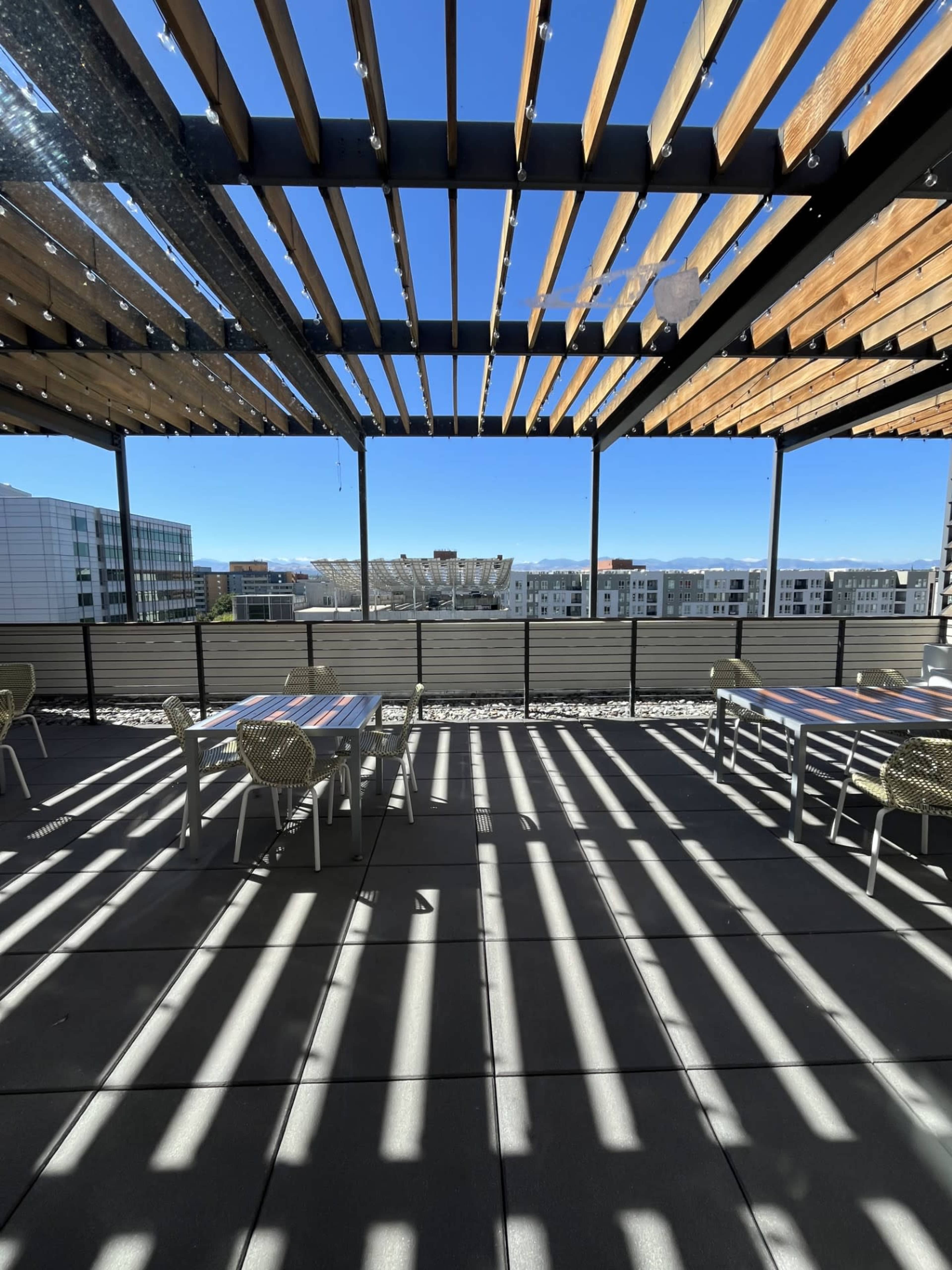 The image shows a rooftop terrace with wooden beams casting shadows on the tile floor, overlooking a cityscape.