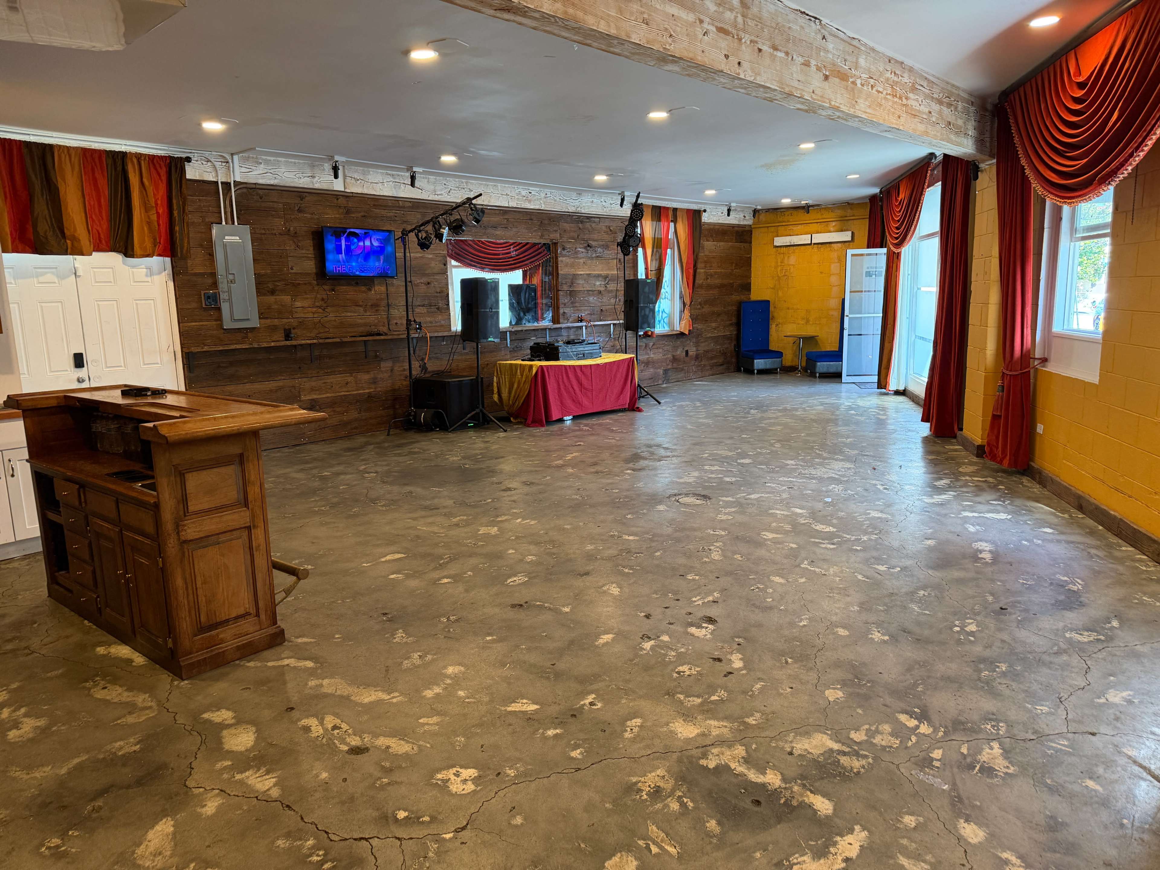 The image shows a spacious room with a polished concrete floor, wooden walls, and colorful curtains, featuring a bar area and a stage setup at one end.