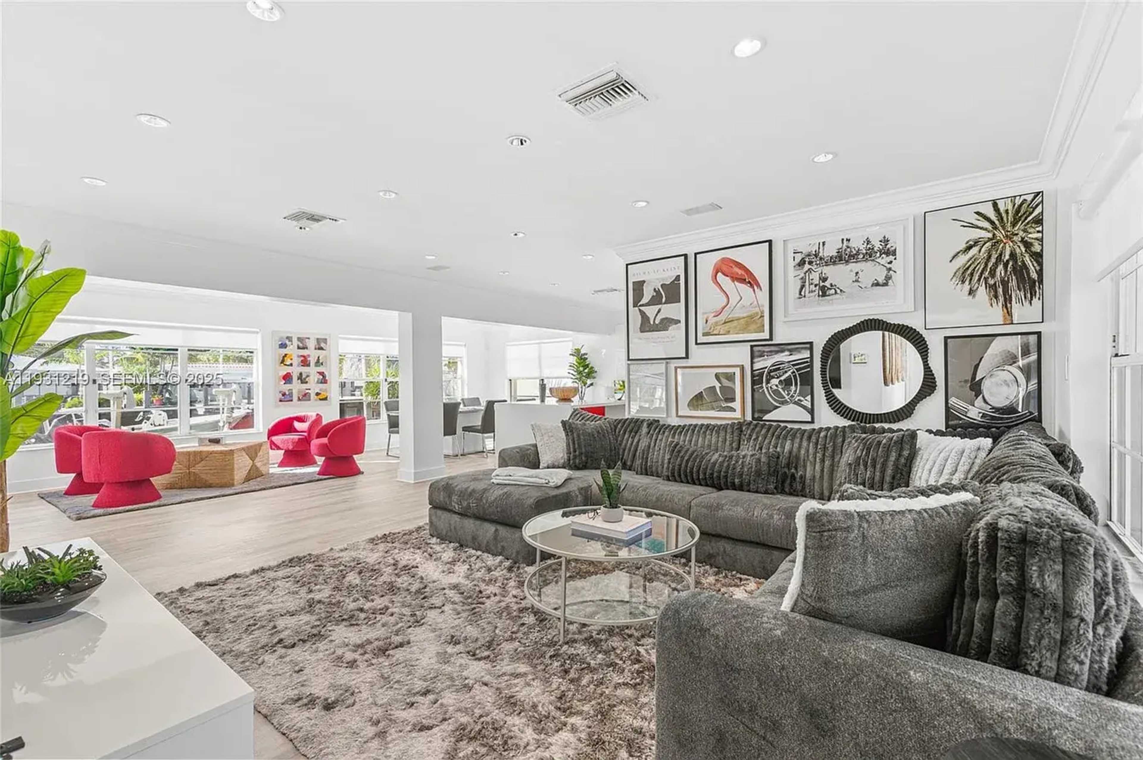 The image shows a spacious, modern living room with a large gray sectional sofa, a round glass coffee table, and various framed artworks on the walls.