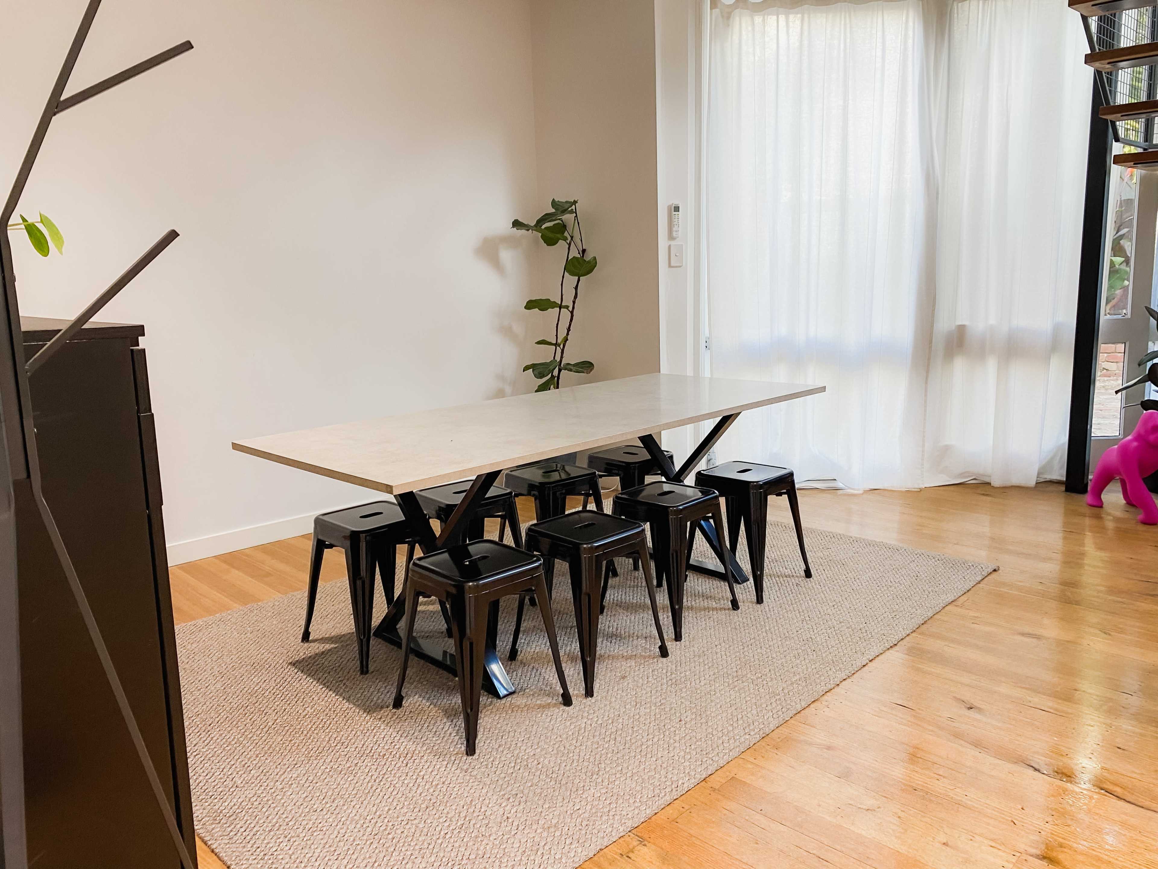 A rectangular table with eight black stools is positioned on a rug in a brightly lit room featuring a large window with flowing curtains.