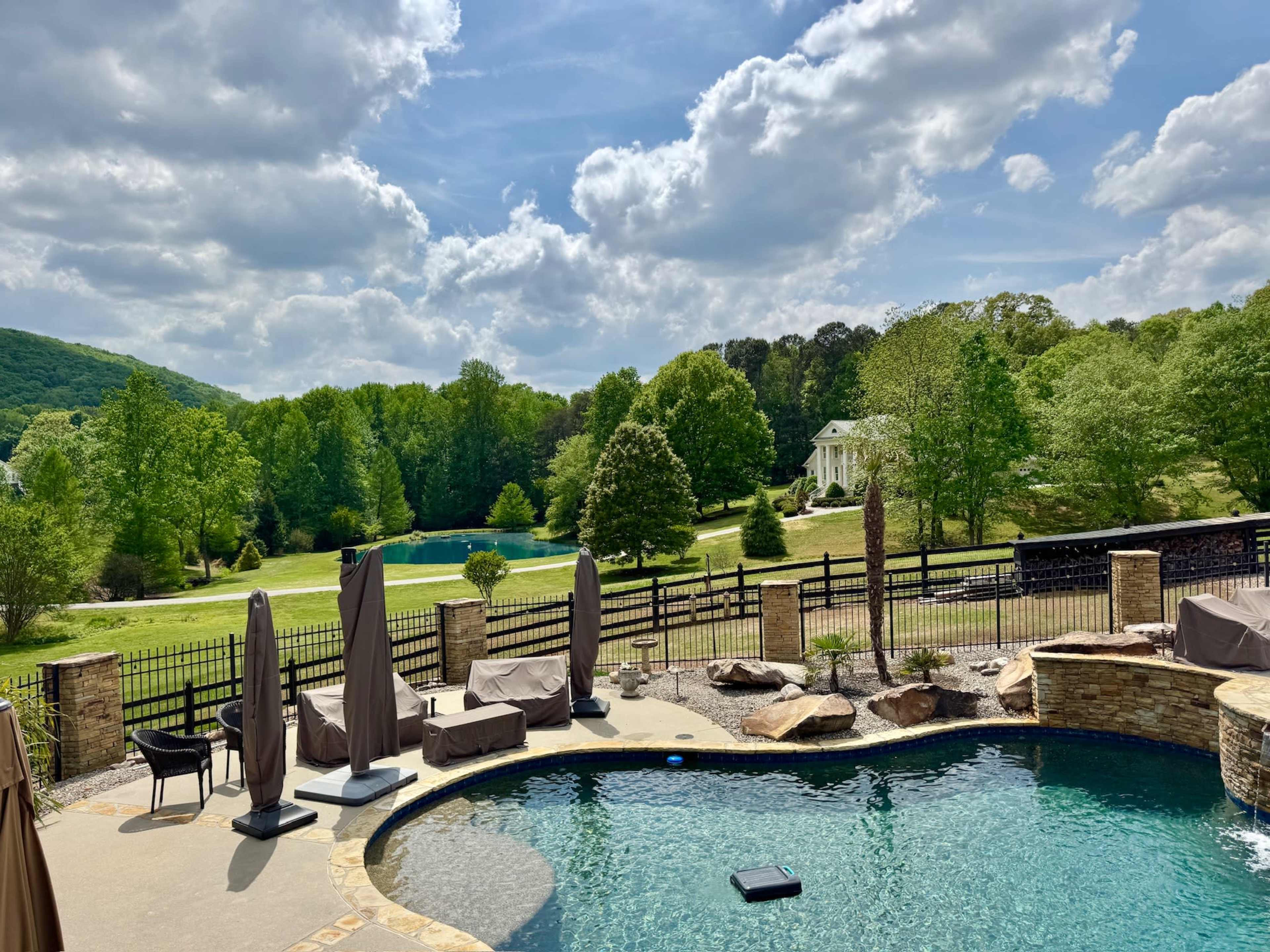The image shows a luxurious outdoor pool area with lounge chairs and umbrellas, overlooking a green landscape and a house in the background.