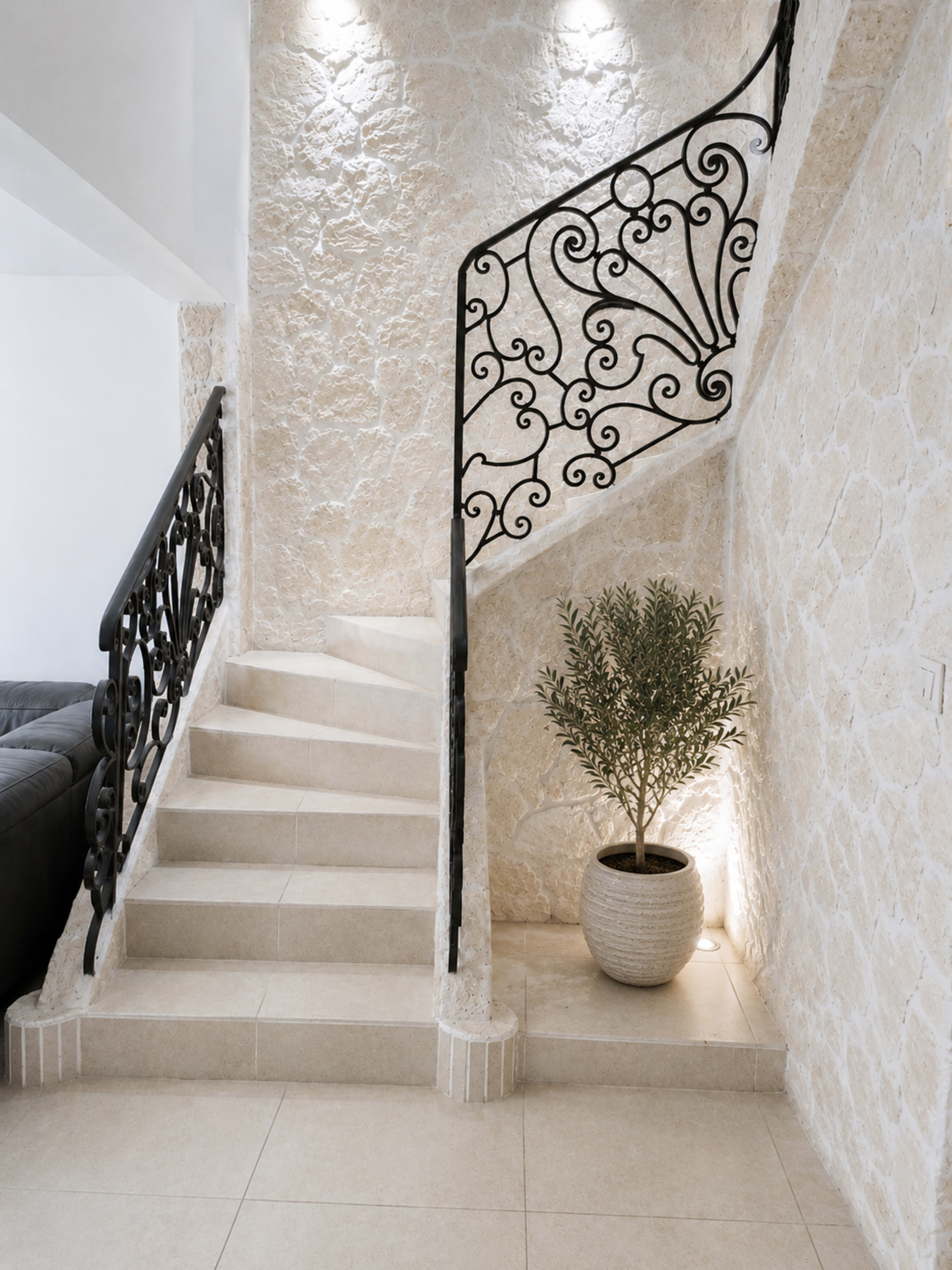 A staircase with a decorative iron railing is located beside a textured stone wall, leading to an upper level, while a potted plant sits in the corner near the base of the stairs.