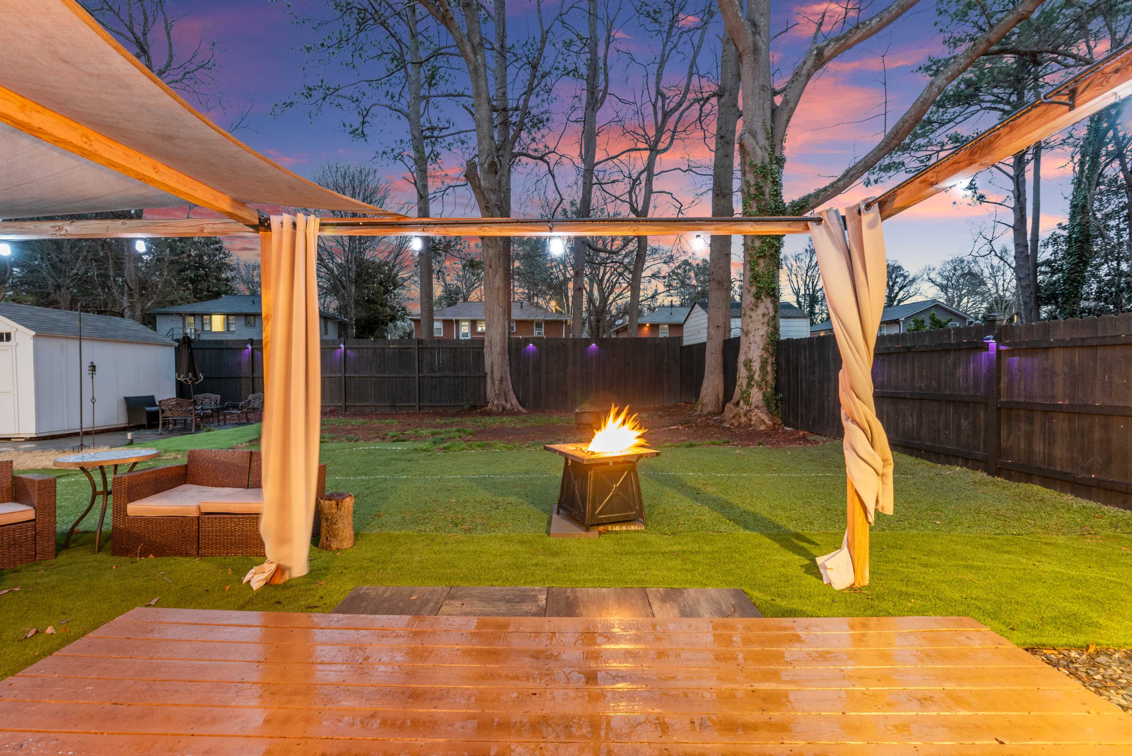 The image shows a backyard with a fire pit surrounded by grass, outdoor seating, and trees, under a colorful sunset sky.