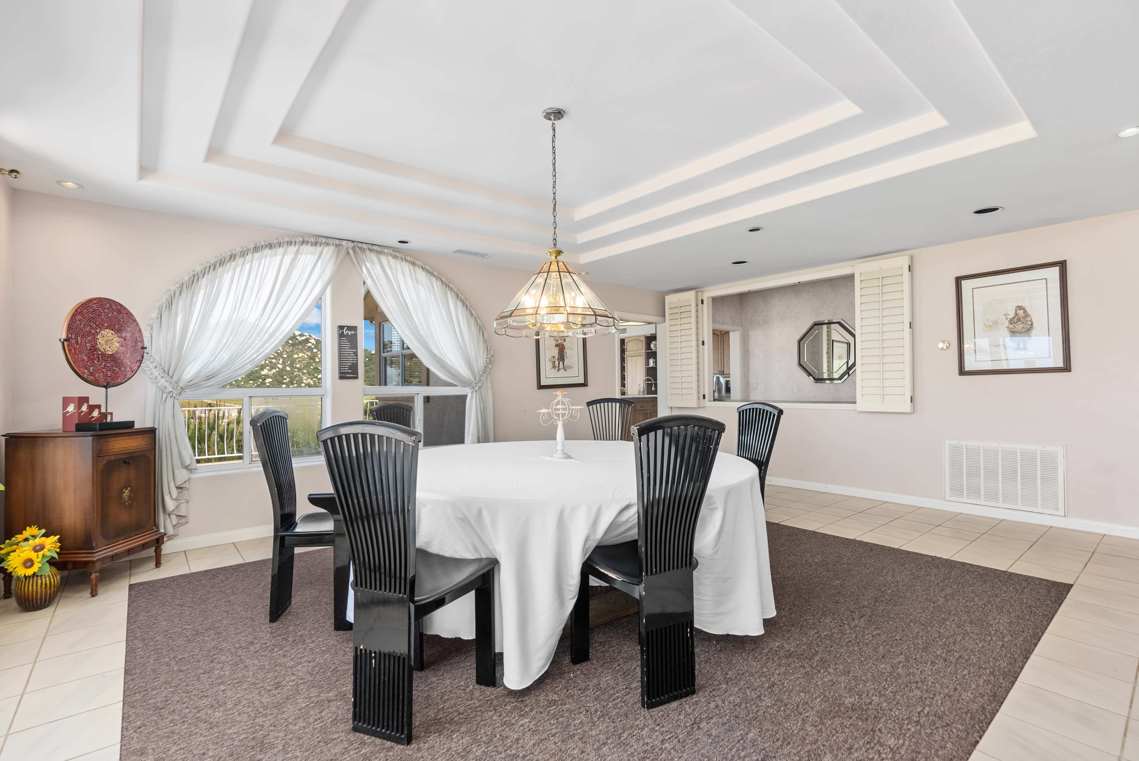 A dining area features a round table with black chairs, surrounded by a light-colored rug, with large windows and decorative elements in the background.