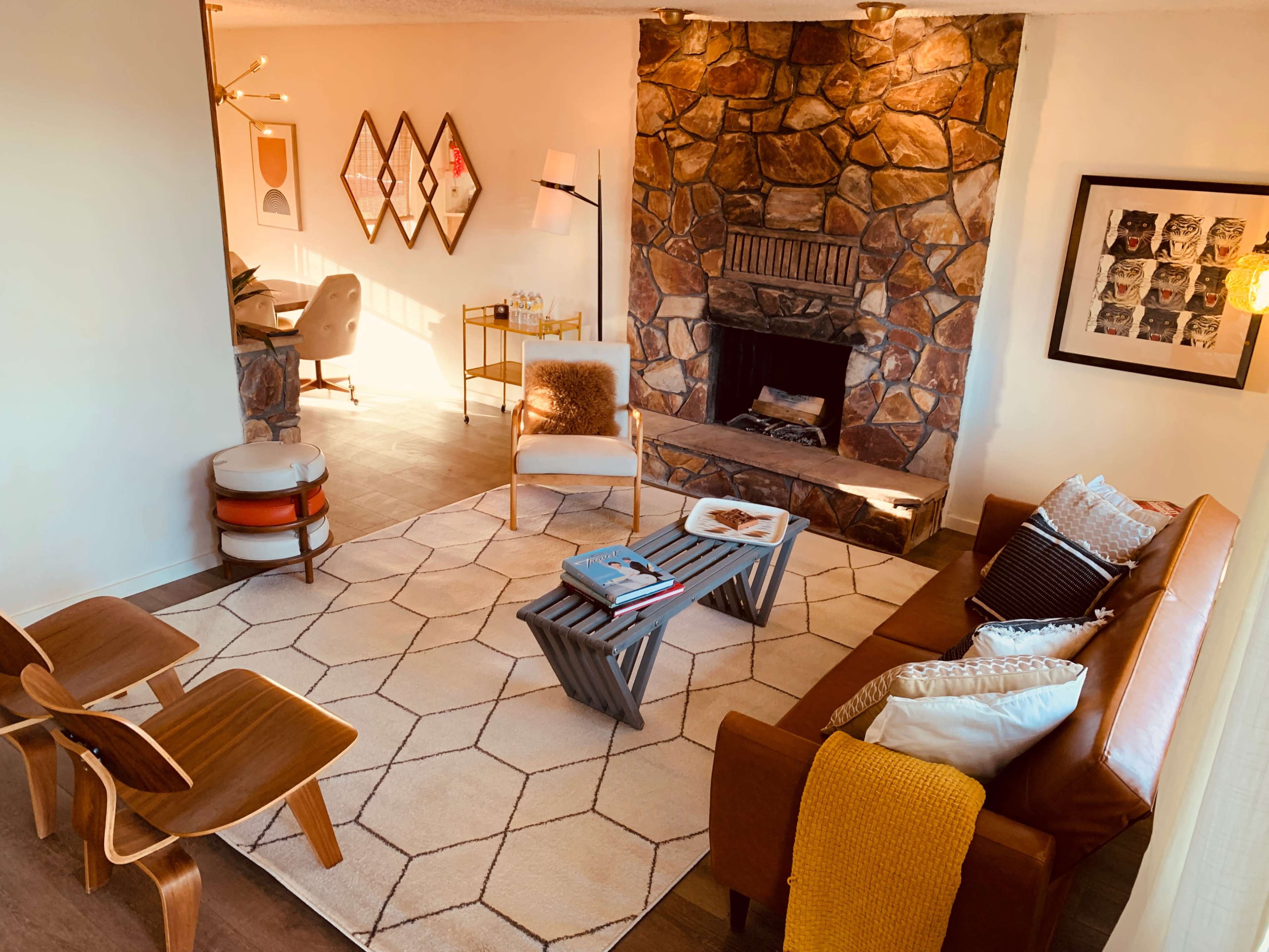 A cozy living room featuring a stone fireplace, a brown leather sofa, a geometric patterned rug, and wooden accent chairs.