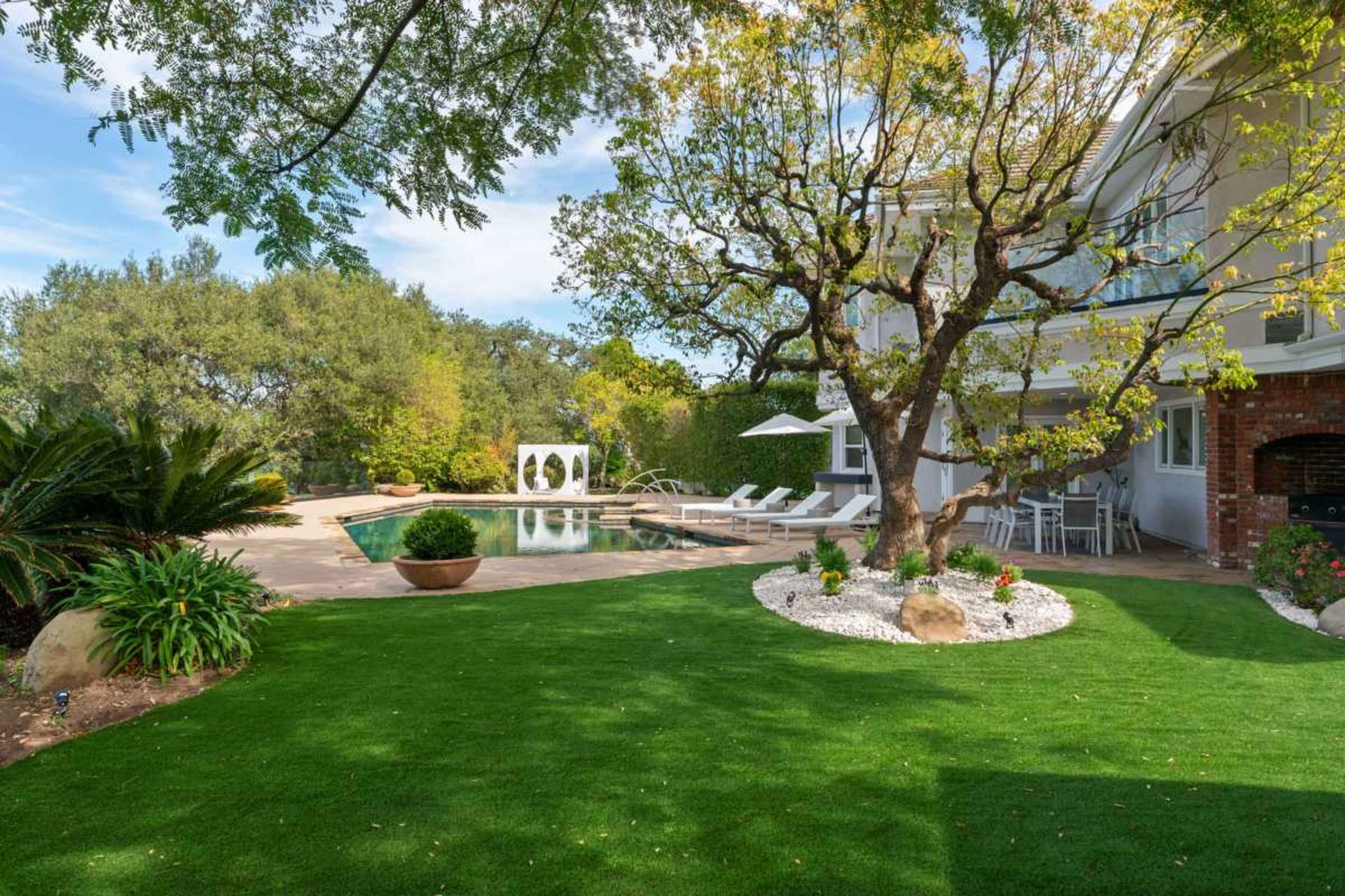 The image shows a landscaped backyard featuring a swimming pool, lounge chairs, a patio area, and a large tree surrounded by greenery.