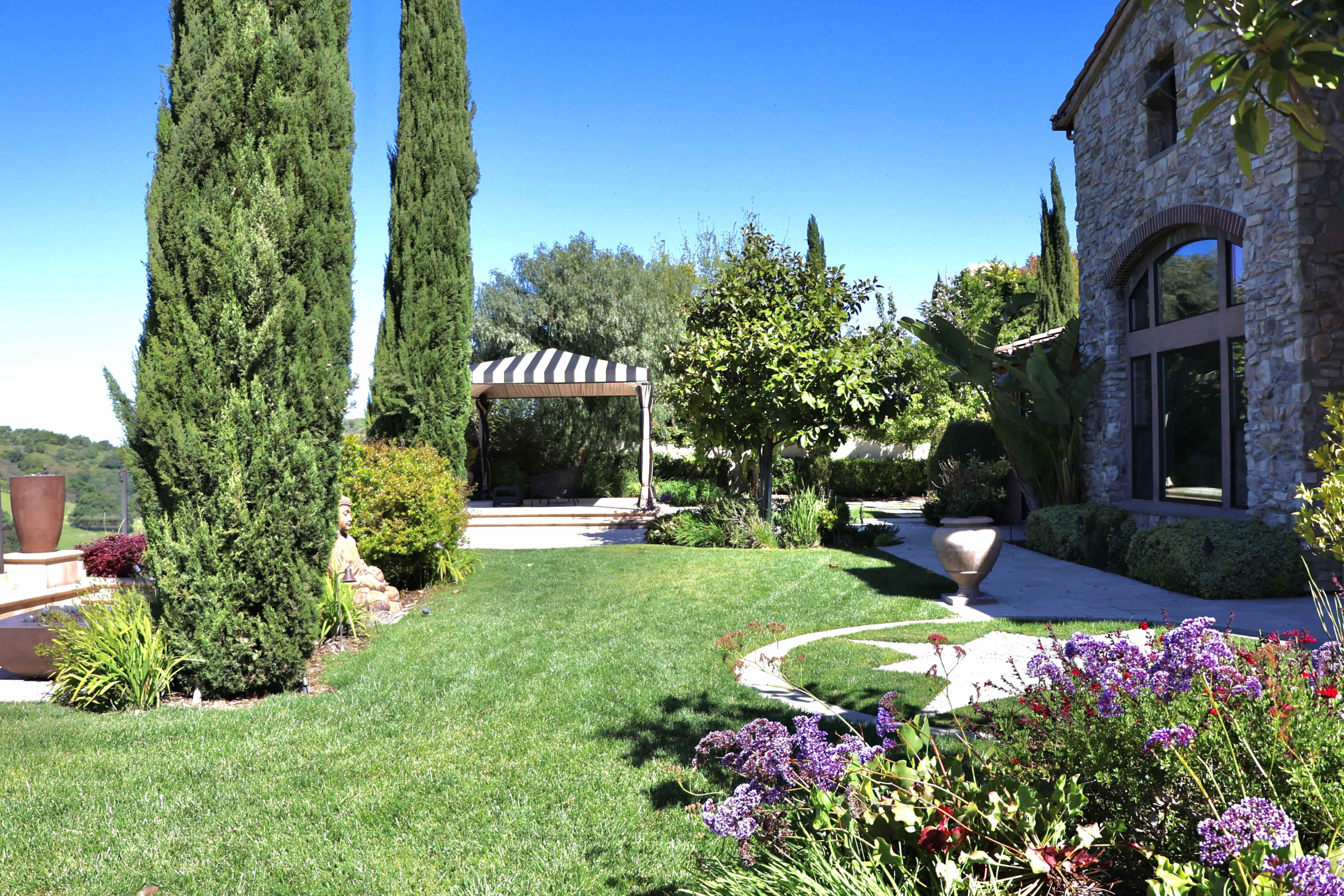 A landscaped garden with neatly trimmed grass, flowering plants, tall cypress trees, and a stone house in the background.