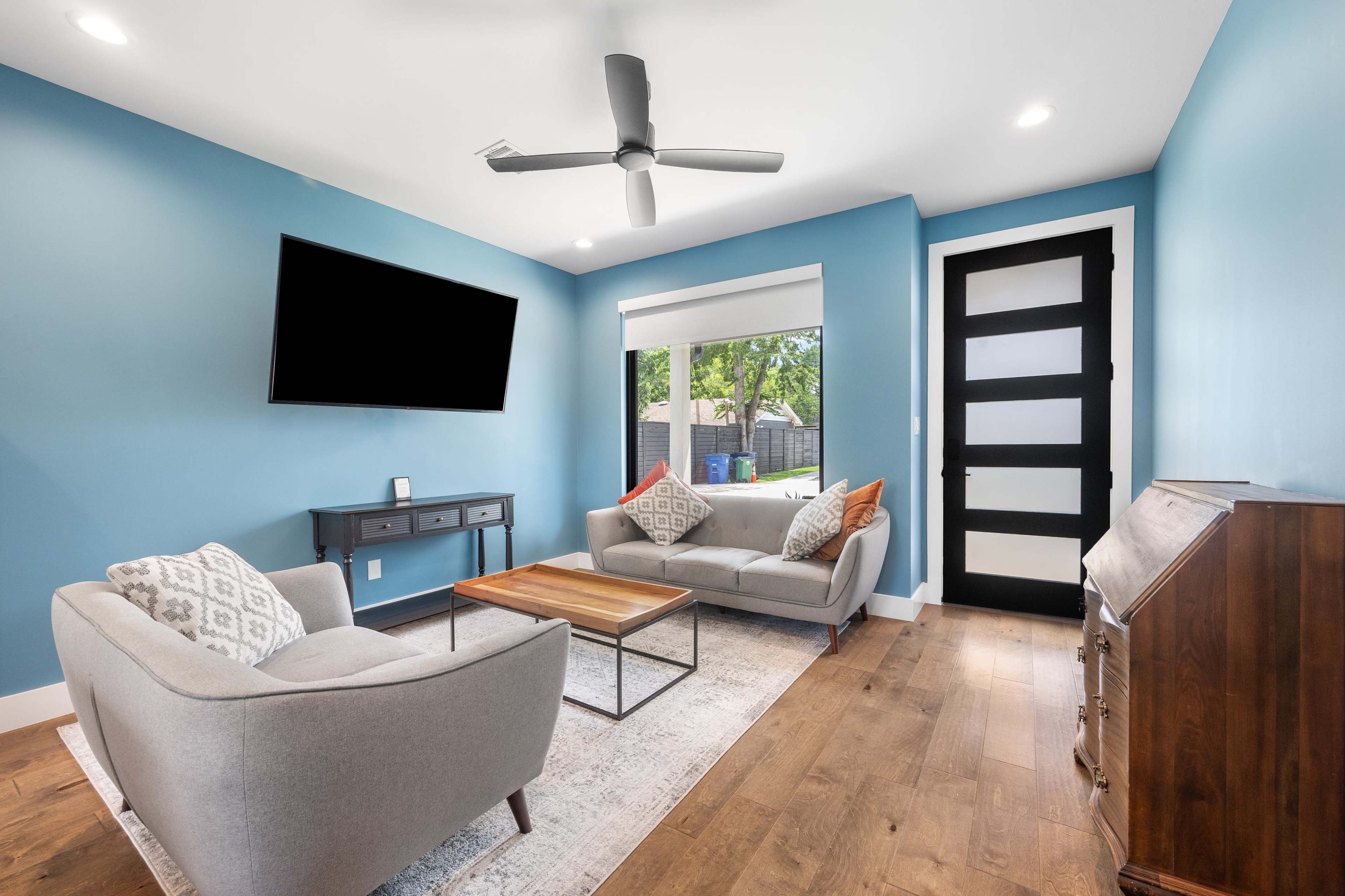 A modern living room features light blue walls, two gray sofas with decorative pillows, a wooden coffee table, and a large window overlooking a green outdoor space.