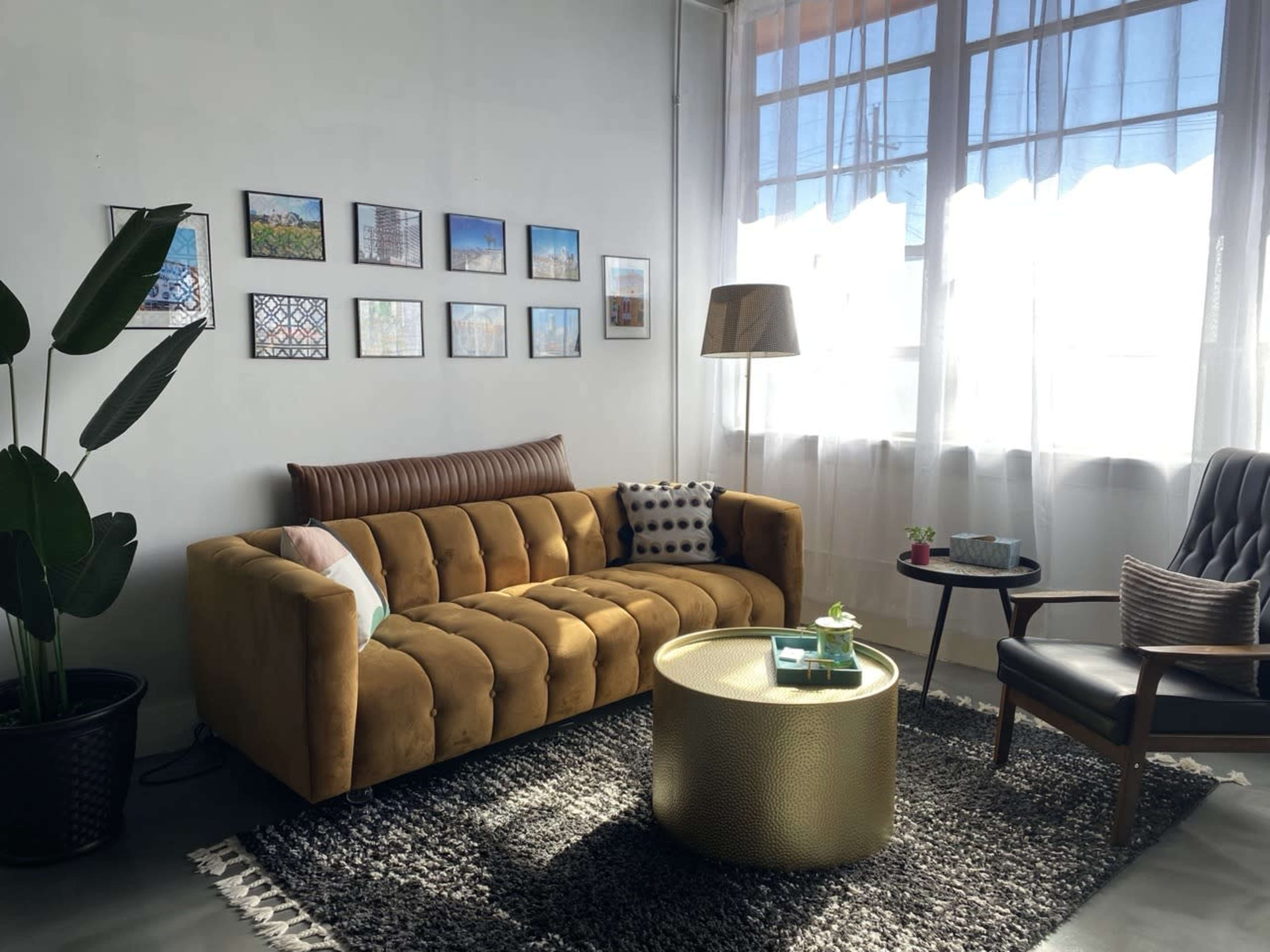 The living room features a mustard-colored velvet sofa, a round gold coffee table, and a black armchair, all set against a backdrop of bright windows and framed artwork on the walls.