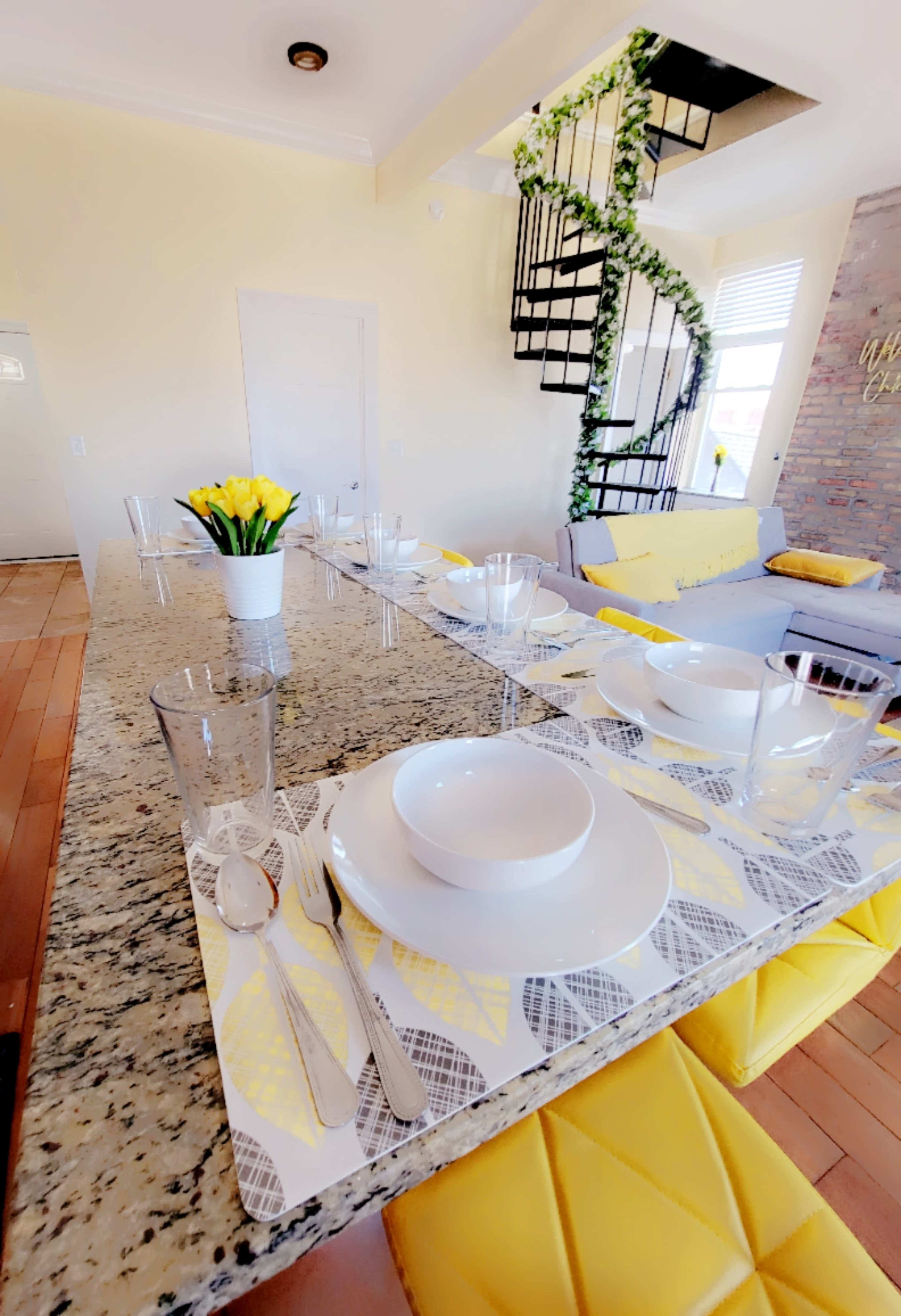 A dining table is set with white plates and glasses on a patterned placemat, alongside a vase of flowers, in a bright room featuring a spiral staircase and a yellow-accented sofa.