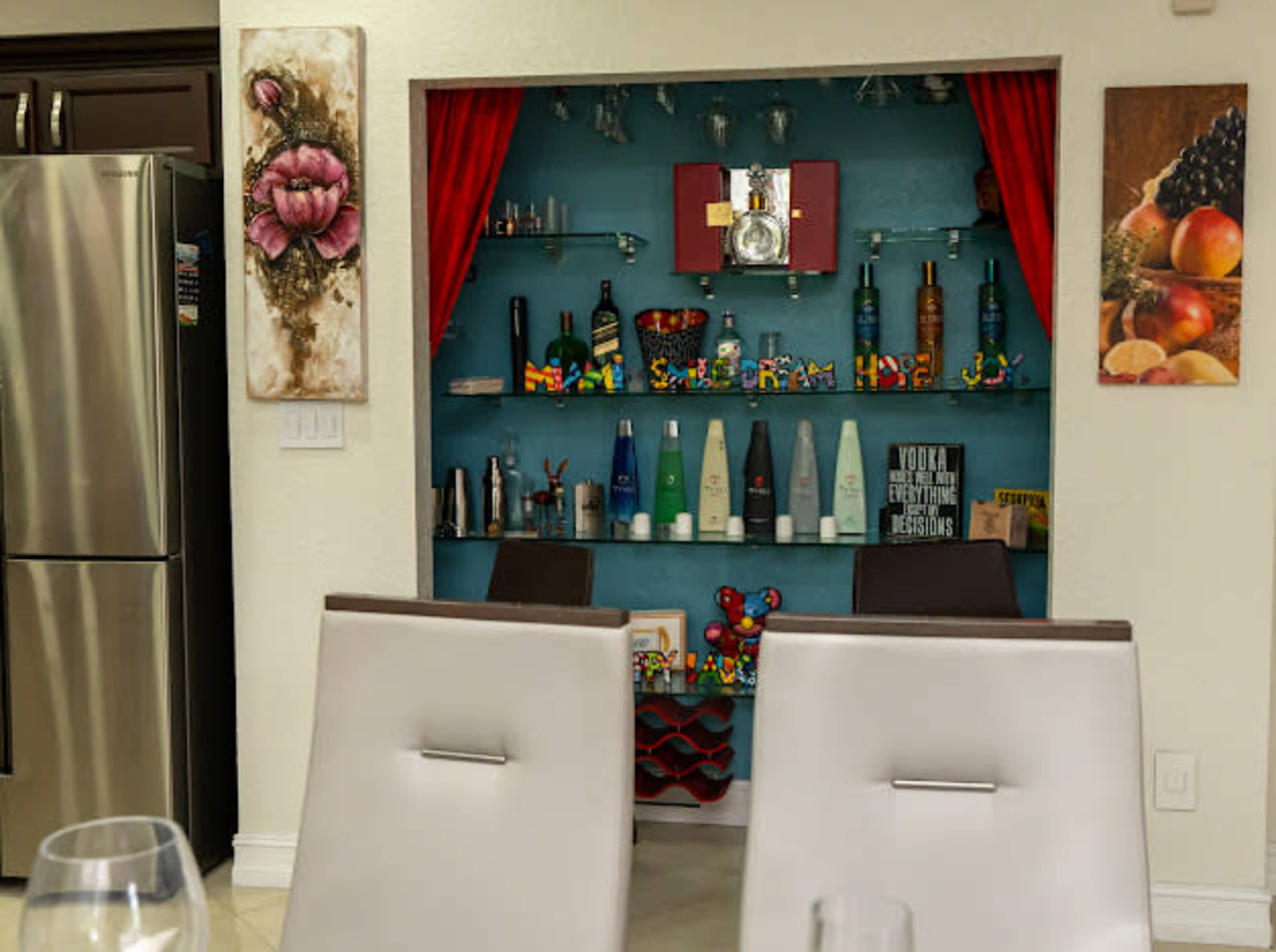 A home bar area with decorative shelves displaying various bottles and colorful toys, framed by red curtains and accompanied by wall art.