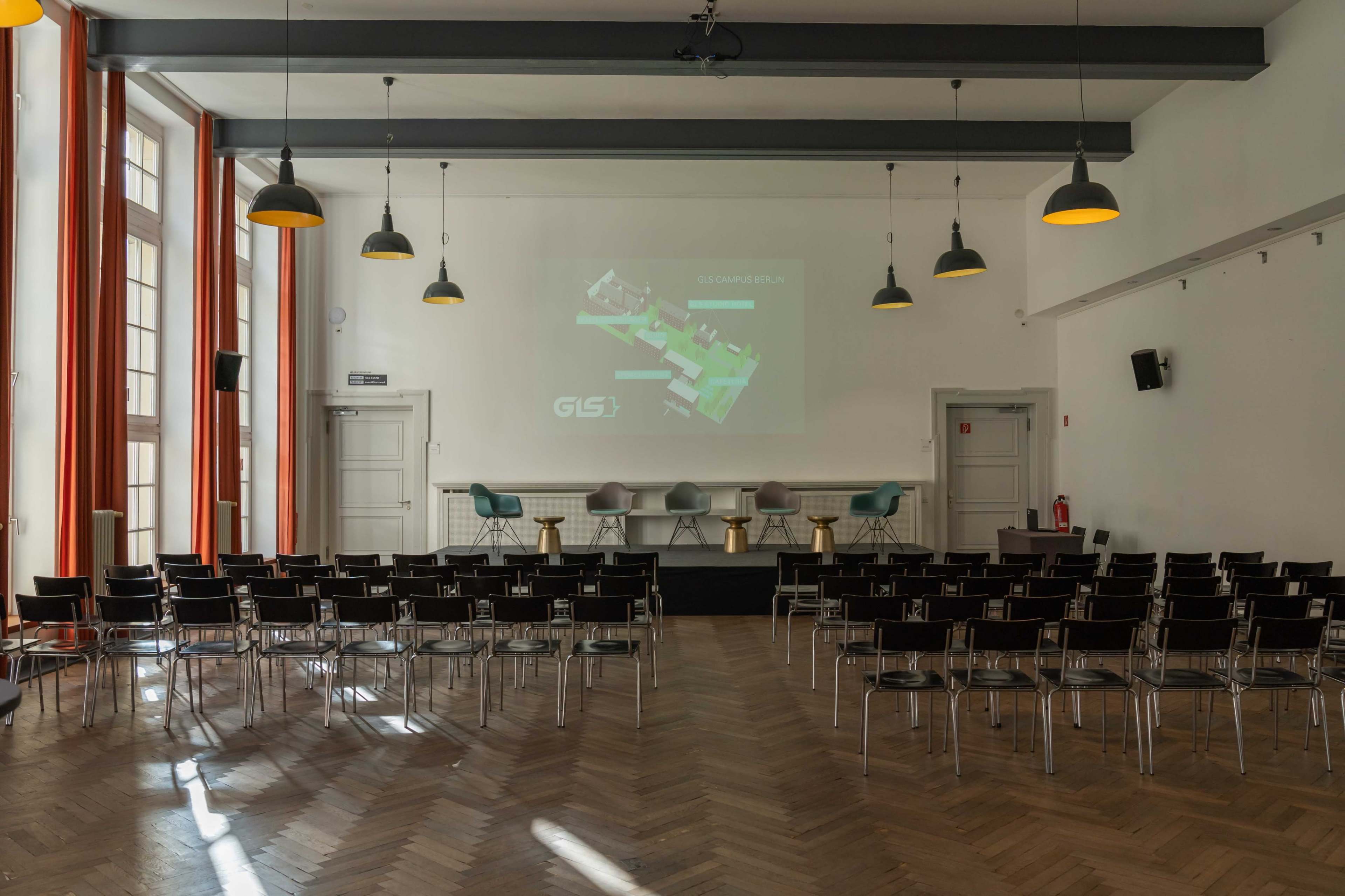 The image shows a conference room setup with rows of chairs facing a projector screen displaying a map, accompanied by a small stage with stools.