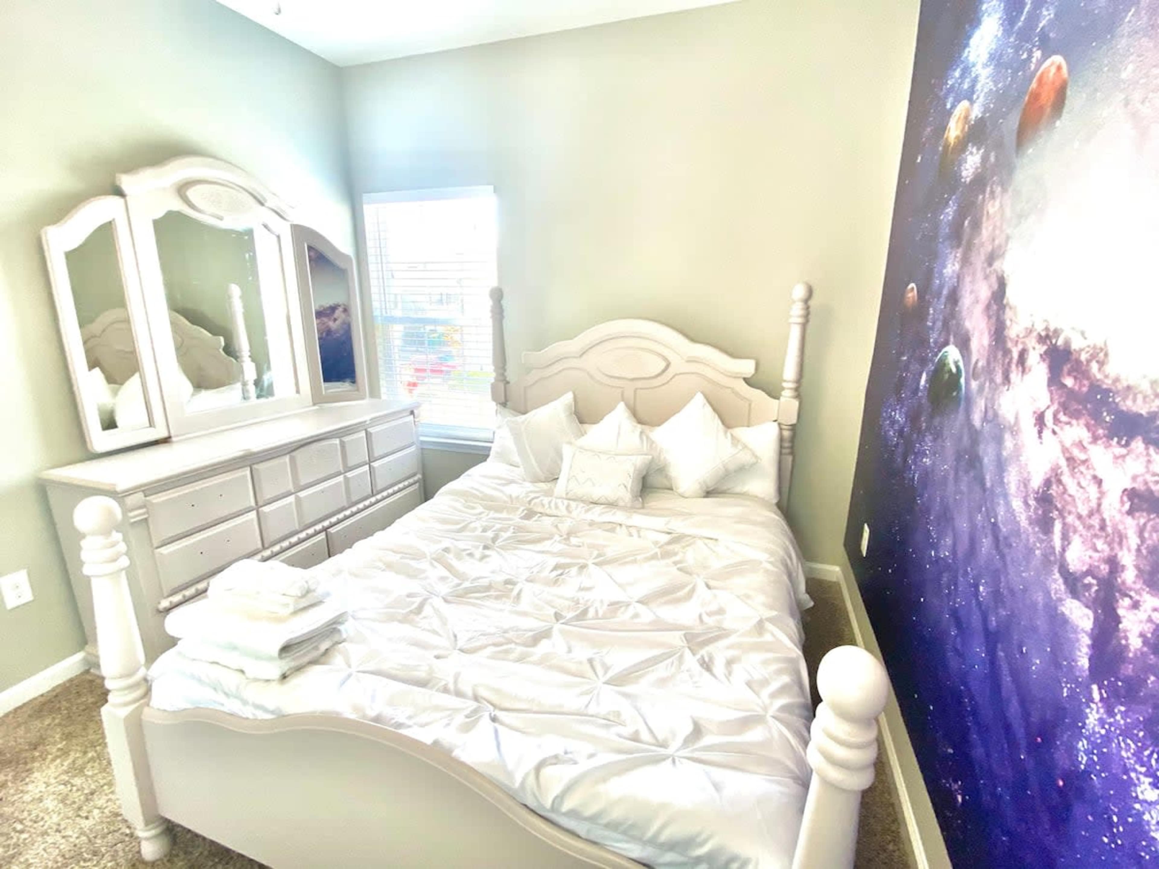 A neatly arranged bedroom with a light-colored wooden bed, a mirrored dresser, and a large wall mural depicting a cosmic scene.