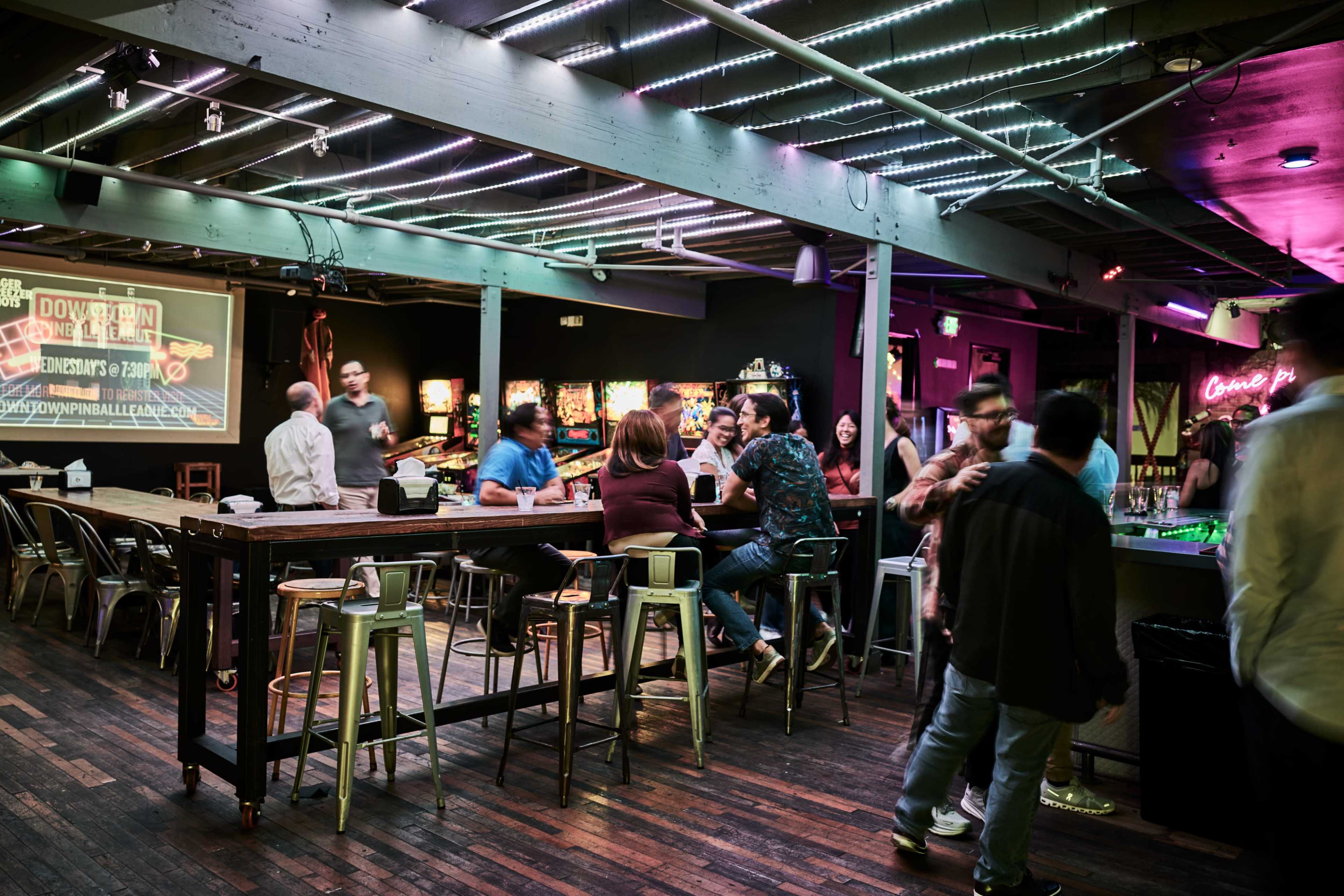 A lively bar scene features people socializing around a high wooden table with illuminated arcade machines in the background.