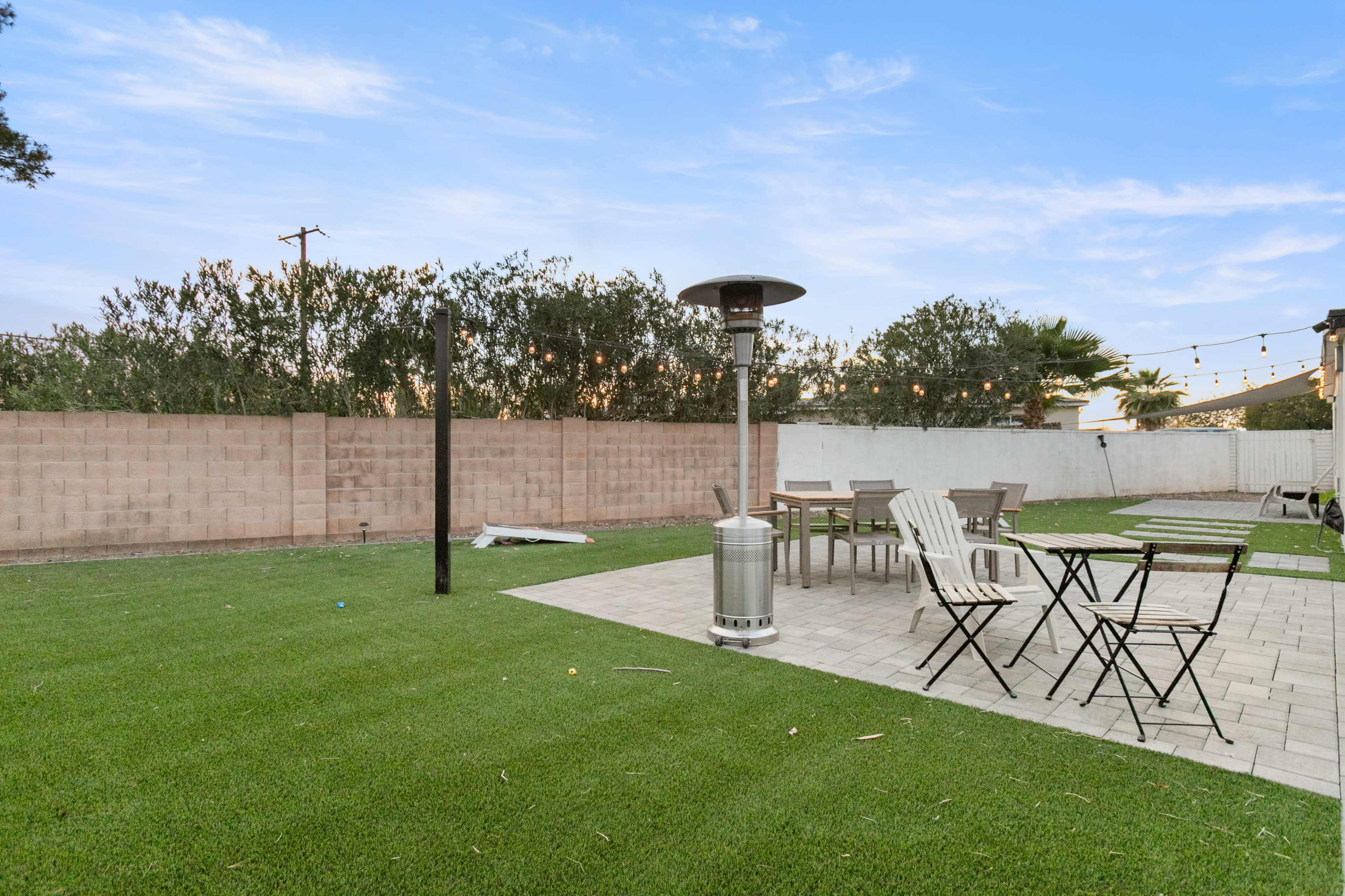 The image shows a spacious backyard with artificial grass, a patio area featuring chairs and tables, and a tall outdoor heater.