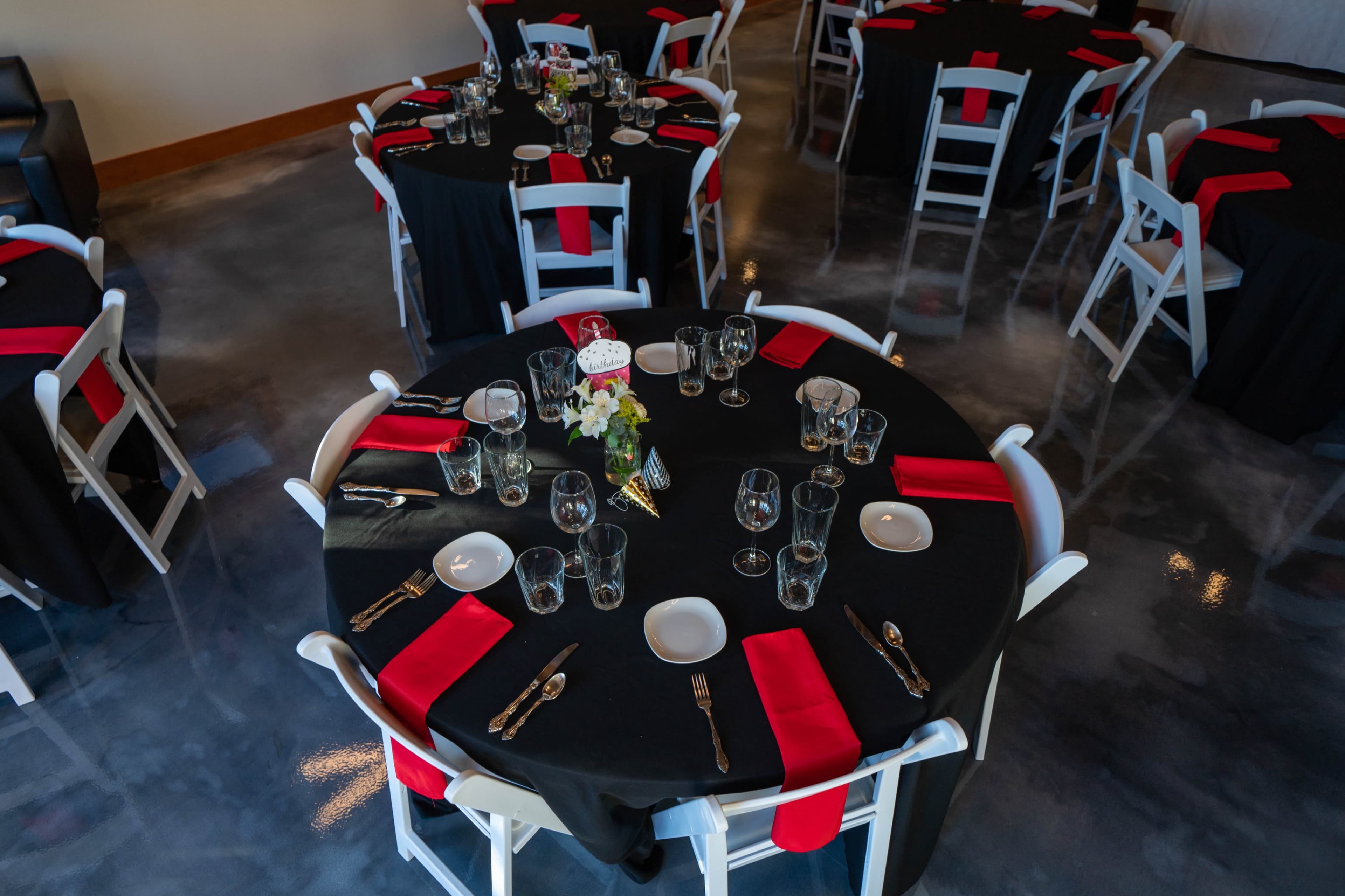 A banquet hall is set up with round tables covered in black cloth, featuring red napkins, glassware, and silverware.