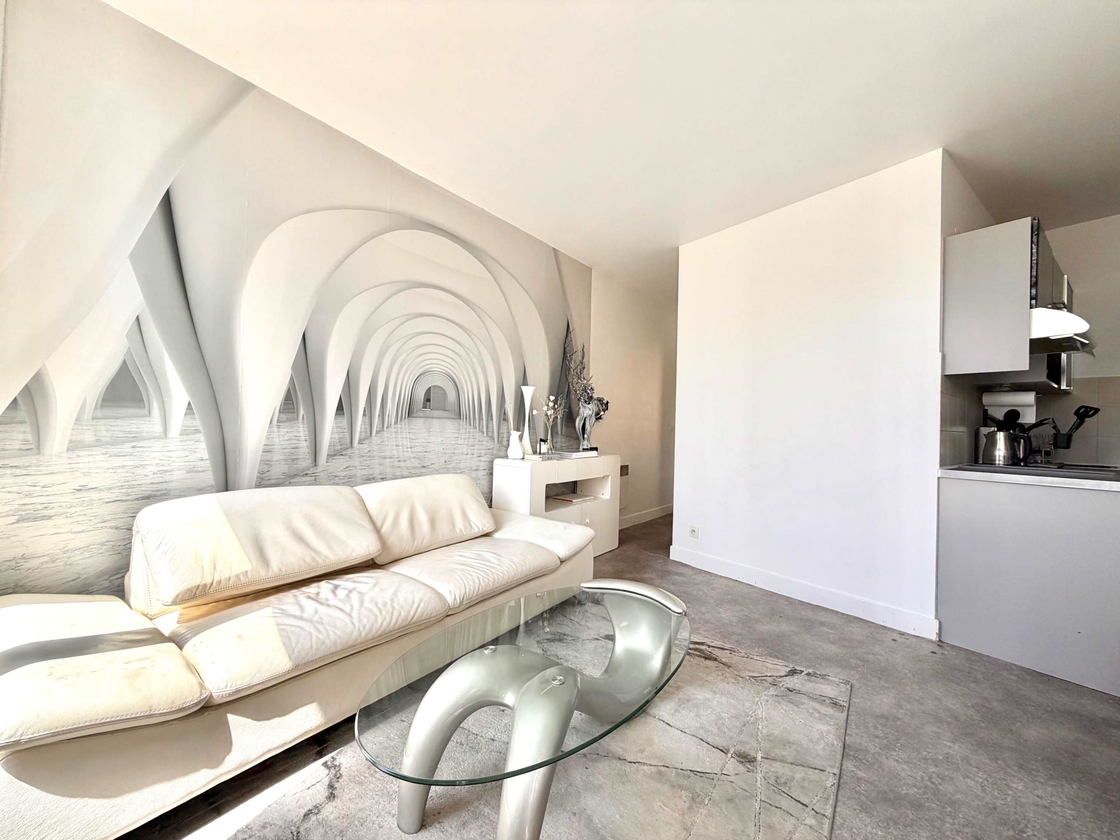The image shows a modern living space featuring a white sofa, a glass coffee table, and a mural of an abstract archway on the wall, with a kitchenette visible in the background.