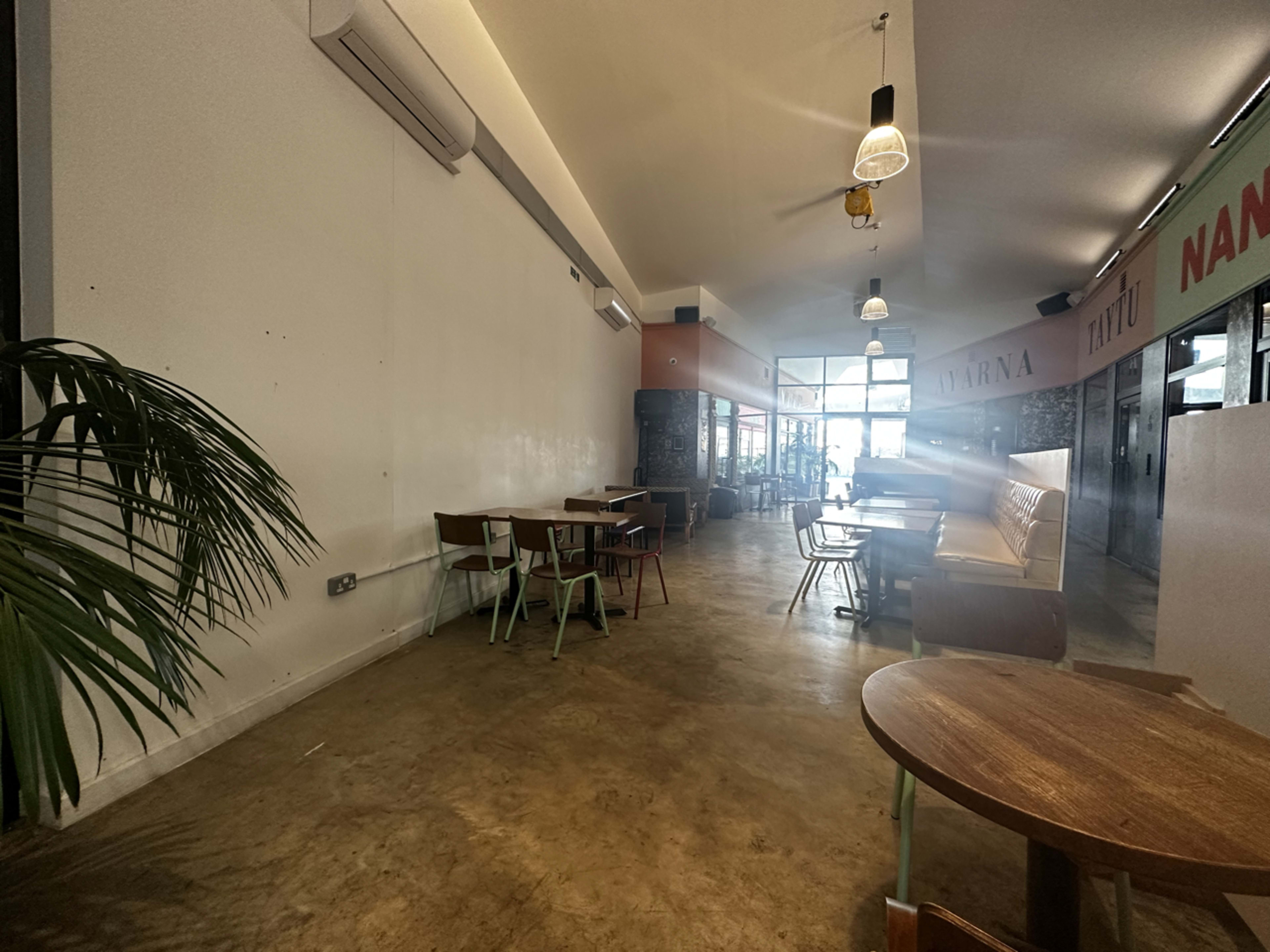 The image shows an empty café interior with a mix of tables and chairs, large windows at the entrance, and a plant in the corner.