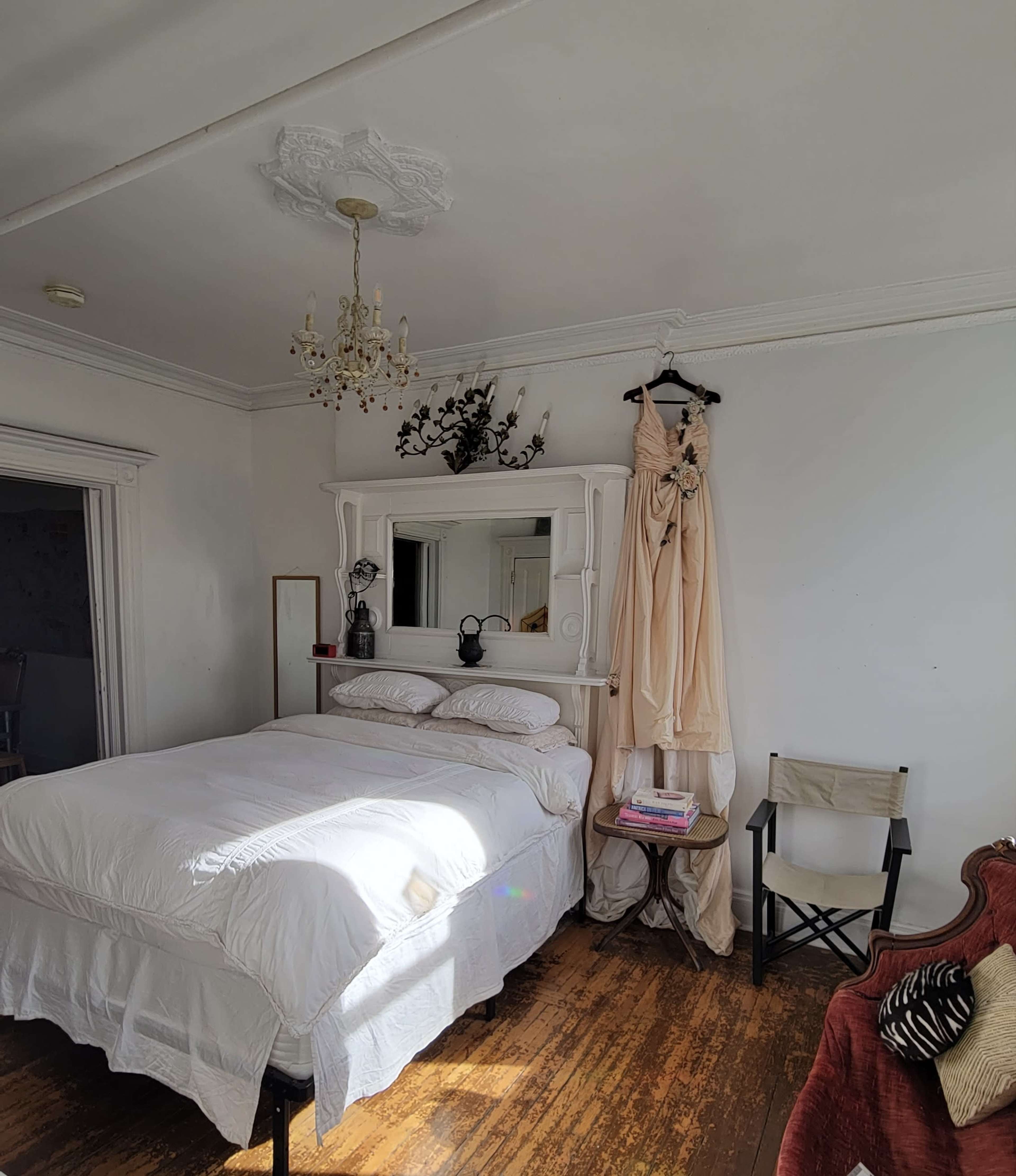 A bedroom features a large bed with white bedding, a mirror above a wooden shelf, and a cream-colored dress hanging beside a chair.