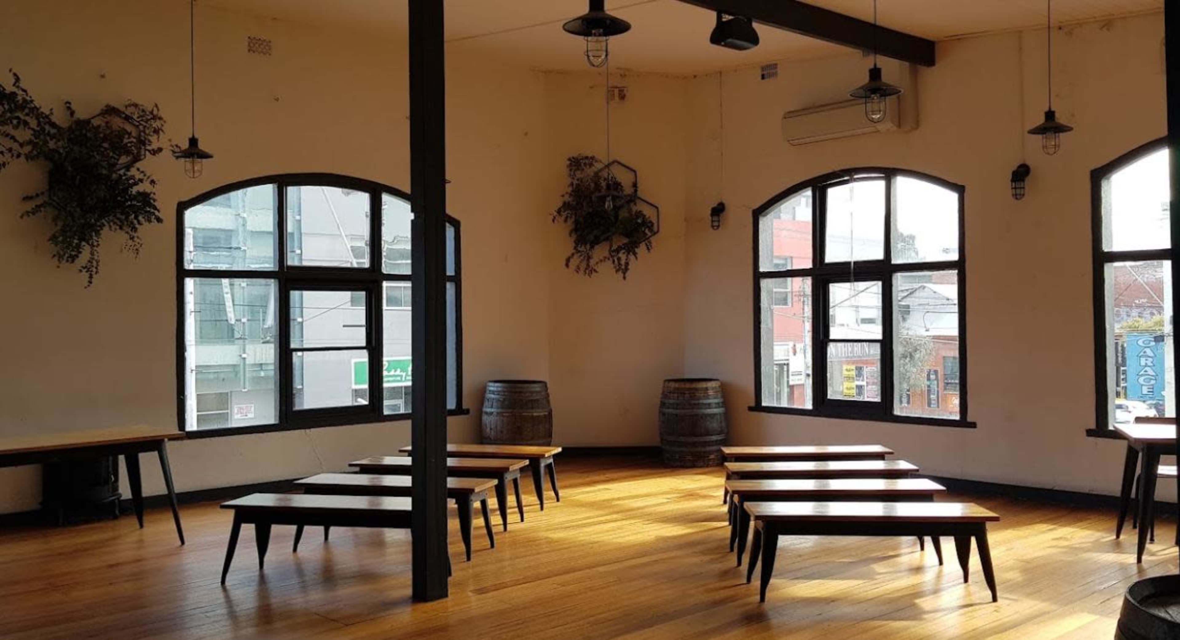 The image shows a spacious room with wooden floors, long tables arranged in rows, large windows allowing natural light, and barrels positioned in the corner.