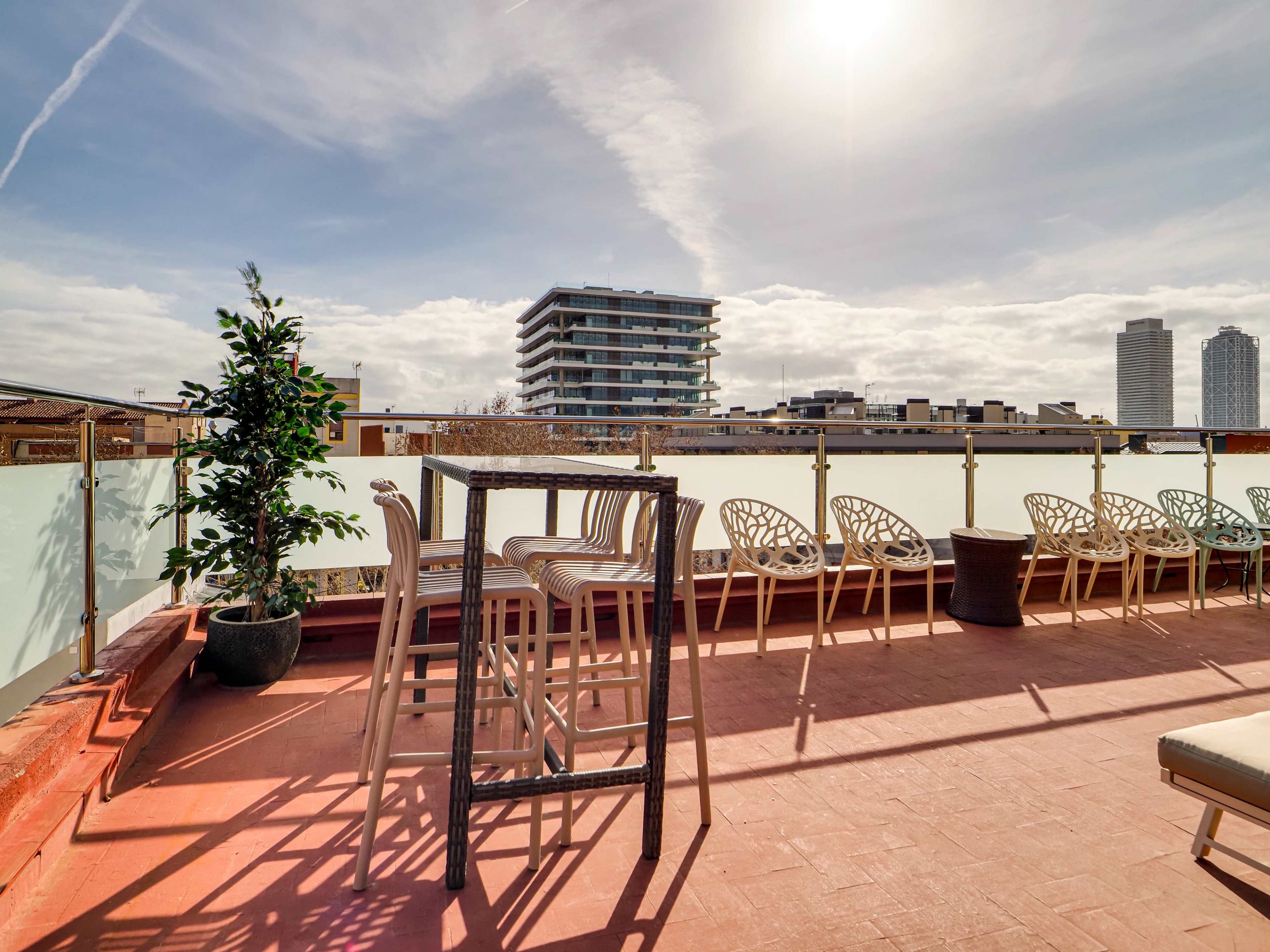 A rooftop terrace features bar stools and decorative plants with a backdrop of modern buildings under a bright sky.