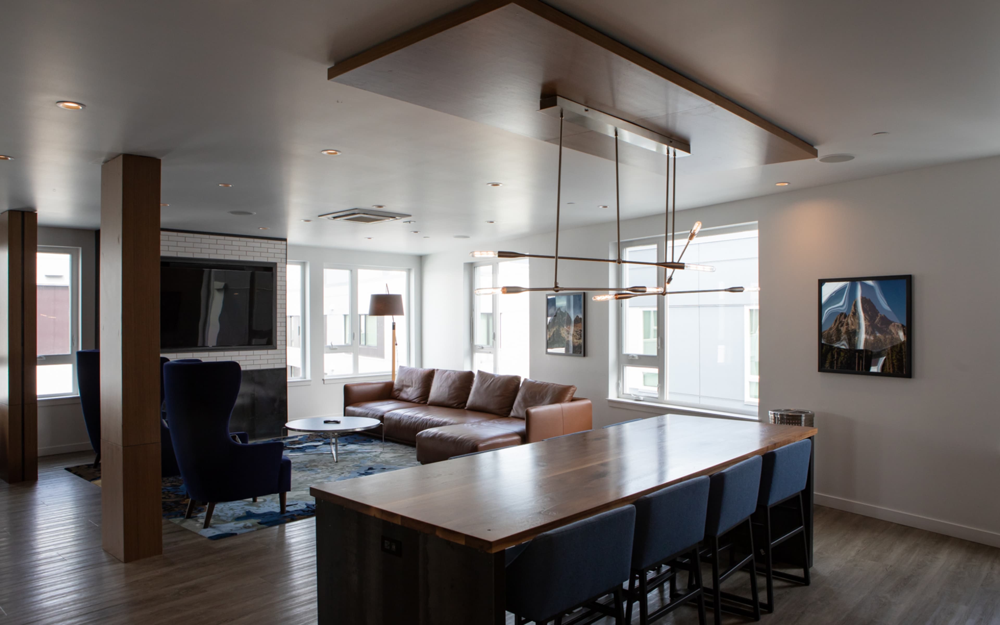 A modern living room with a leather sofa, a dining table with blue chairs, and large windows allowing natural light.