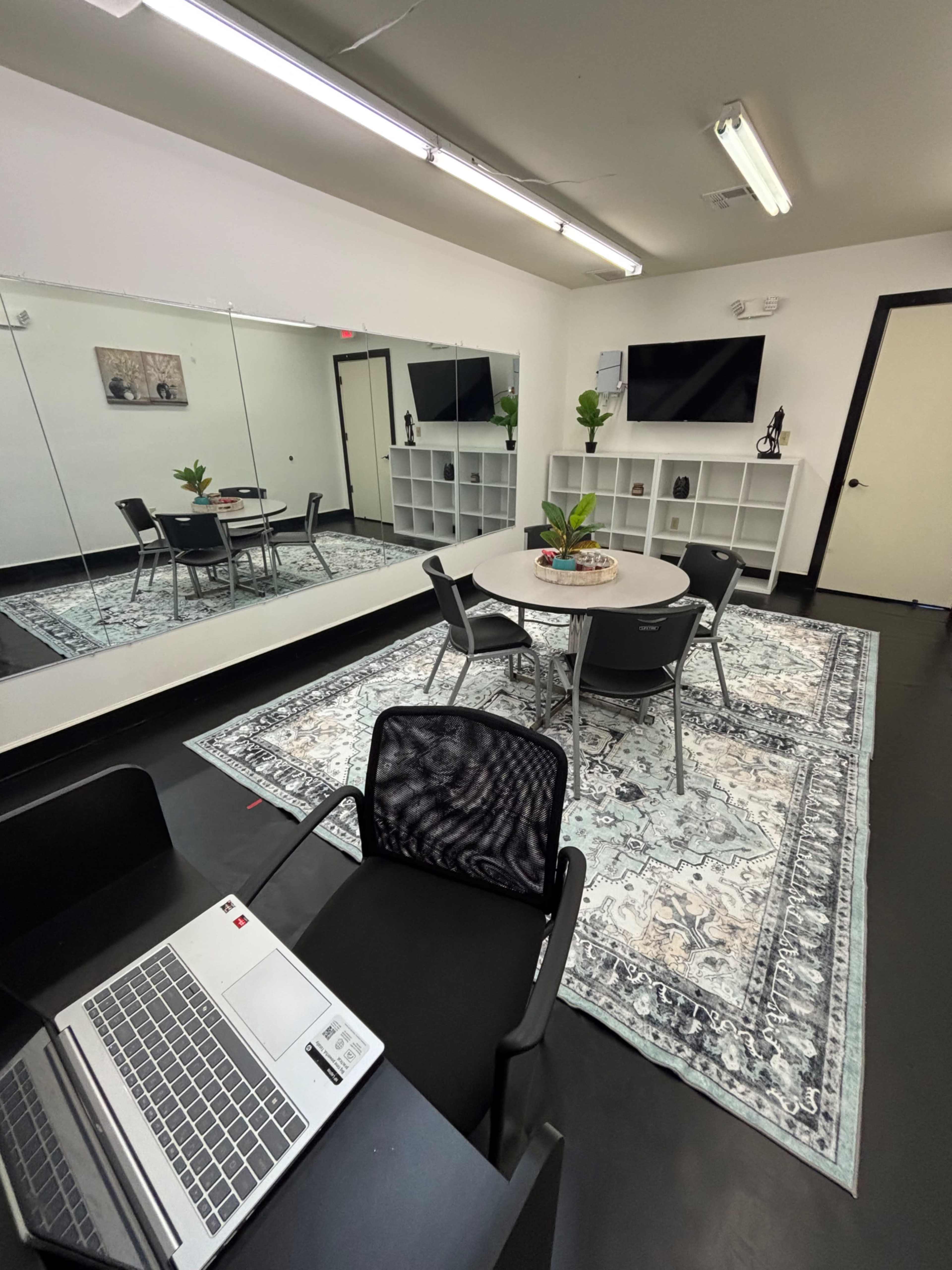 The image shows a modern office space featuring a round table with four chairs, two wall-mounted televisions, a large mirror, and a decorative area rug with plants in the center.