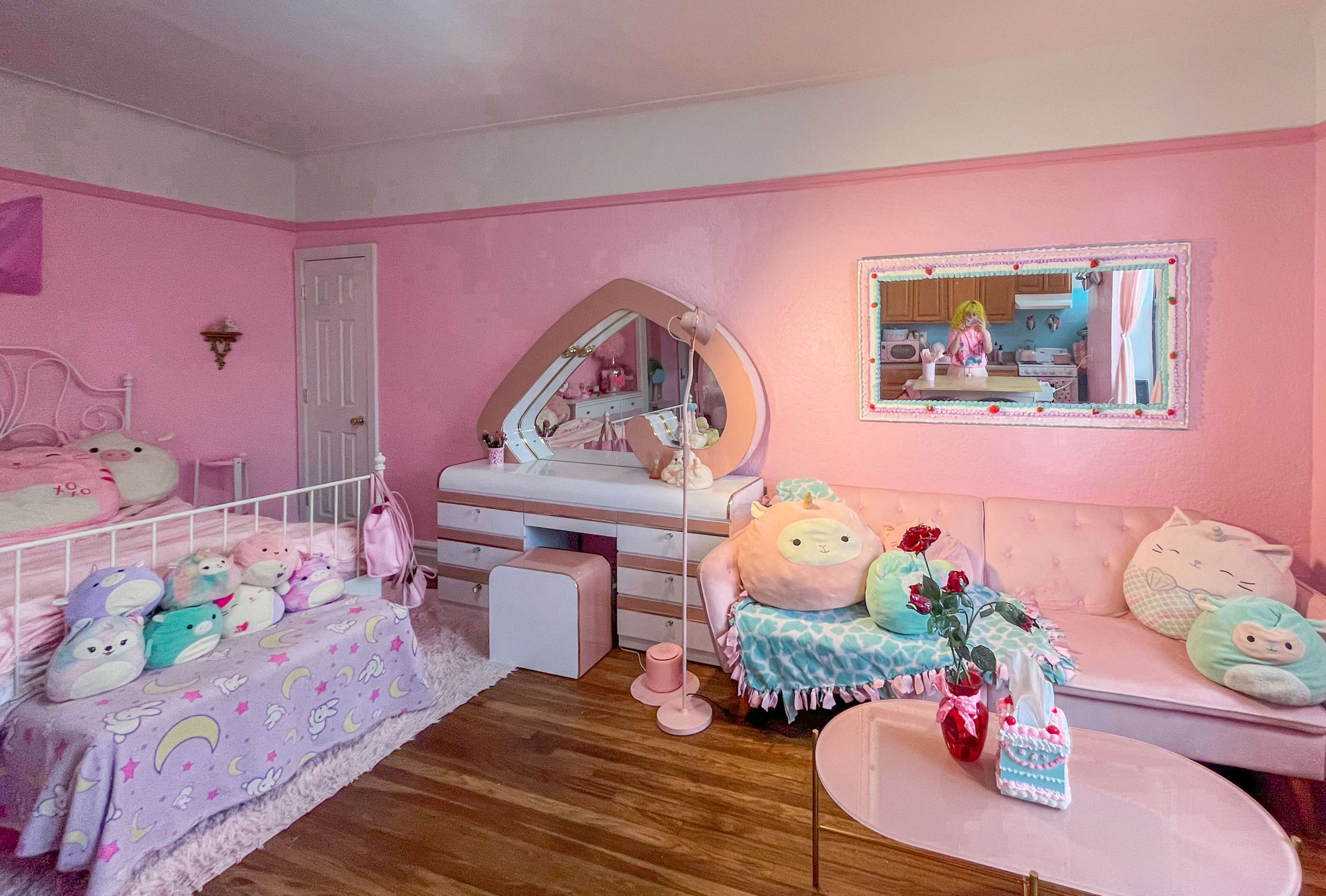The room features pink walls, a bed with a pastel blanket, a mirrored dresser, and a couch adorned with plush toys.
