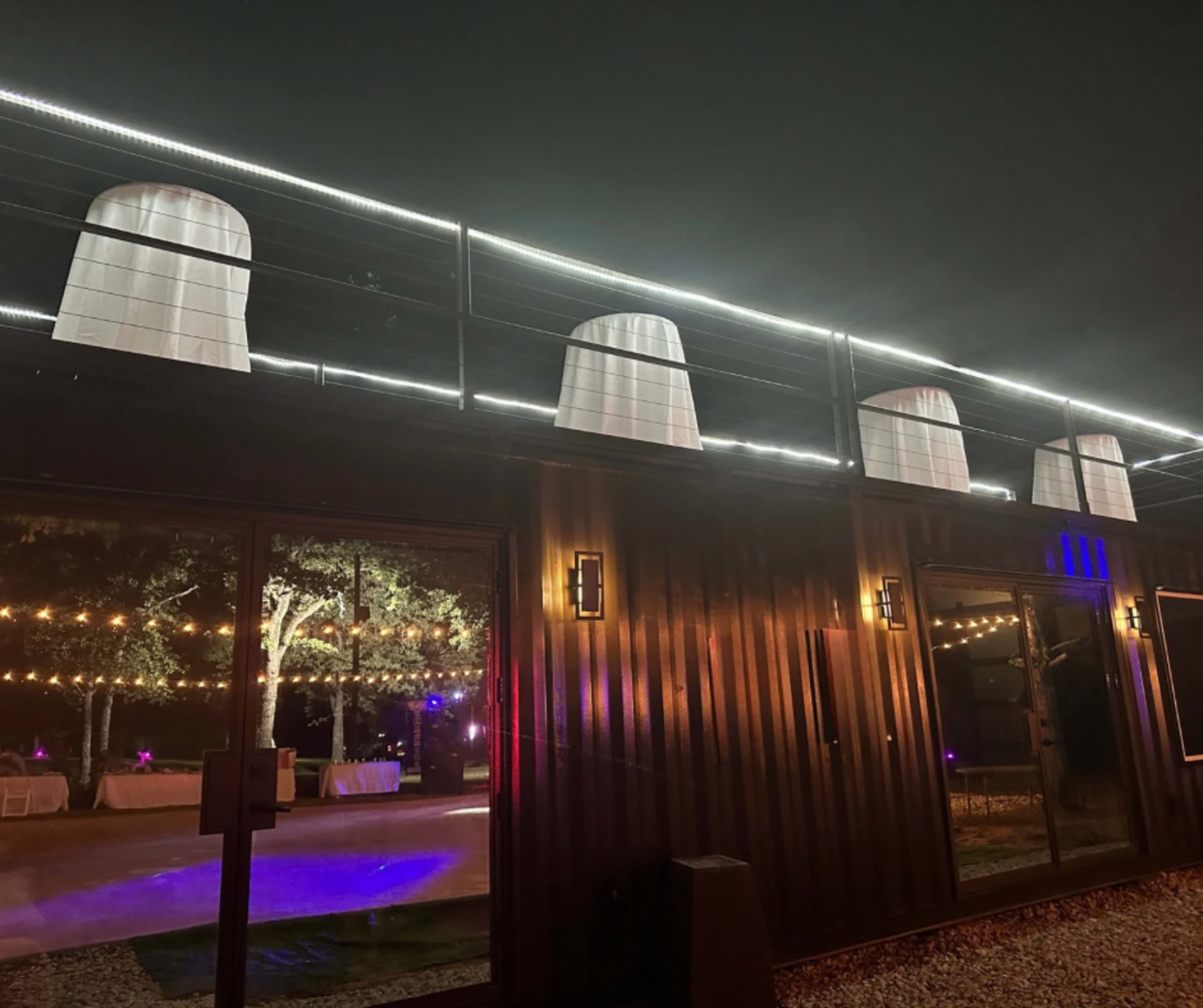 The image shows a modern building with a dark exterior, featuring white draped structures on the roof and illuminated string lights in the background.