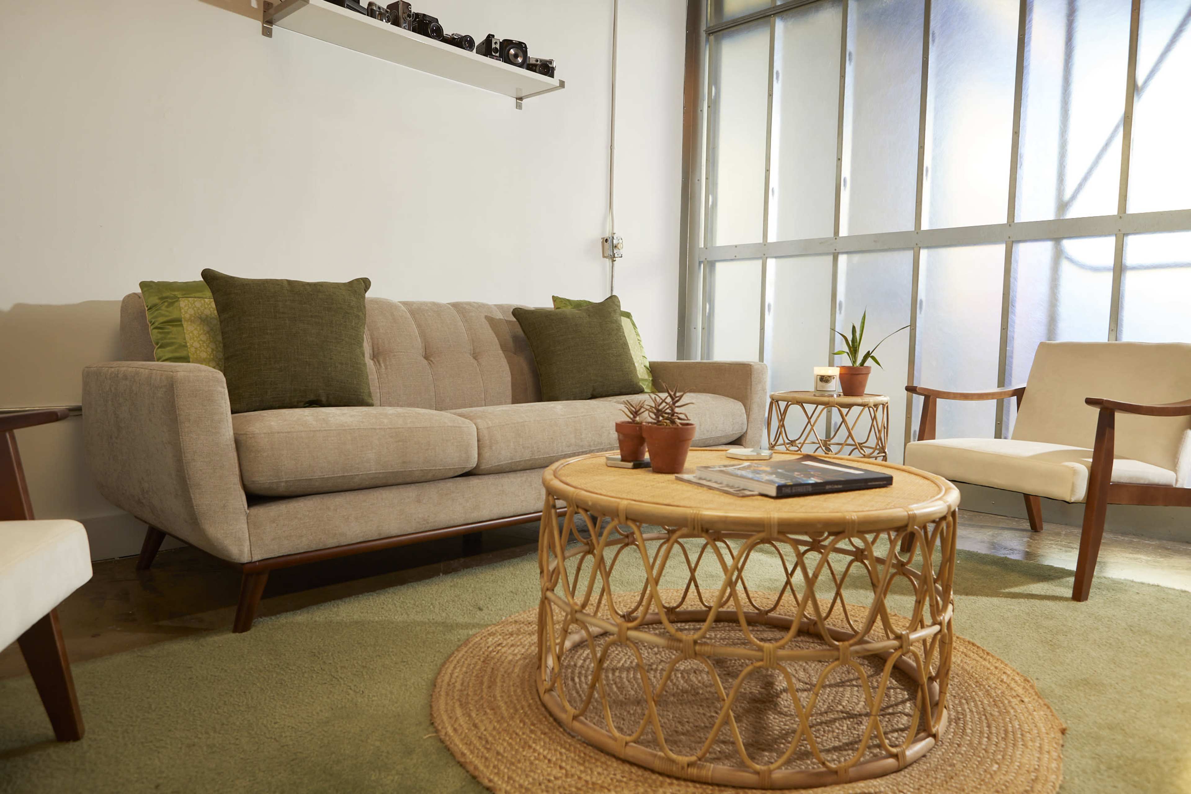 A light-colored couch with green accent pillows is positioned across from a round rattan coffee table on a circular area rug in a well-lit room with large windows.