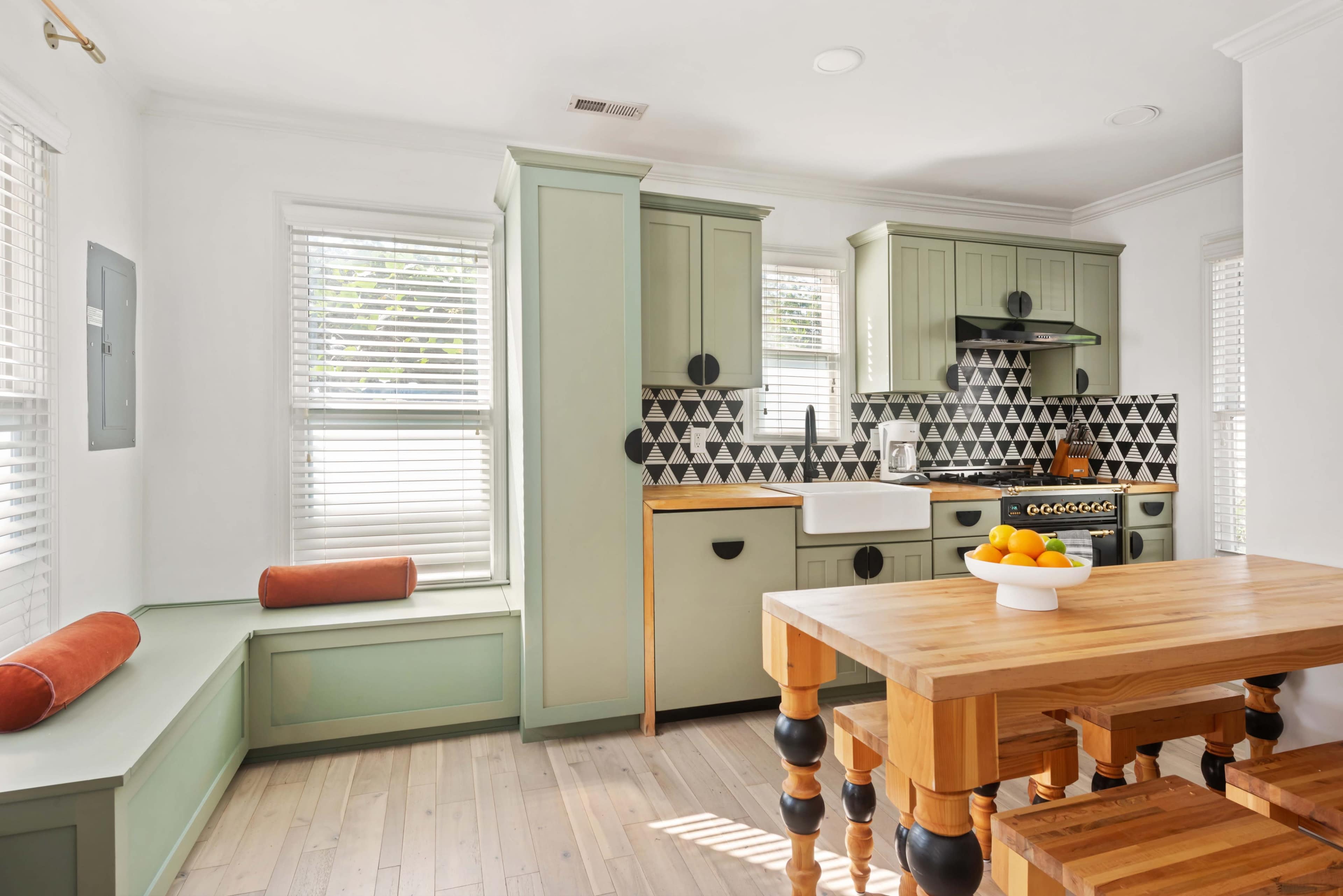 The image shows a modern kitchen featuring light green cabinetry, a patterned black and white backsplash, and a wooden dining table with a bowl of fruit.