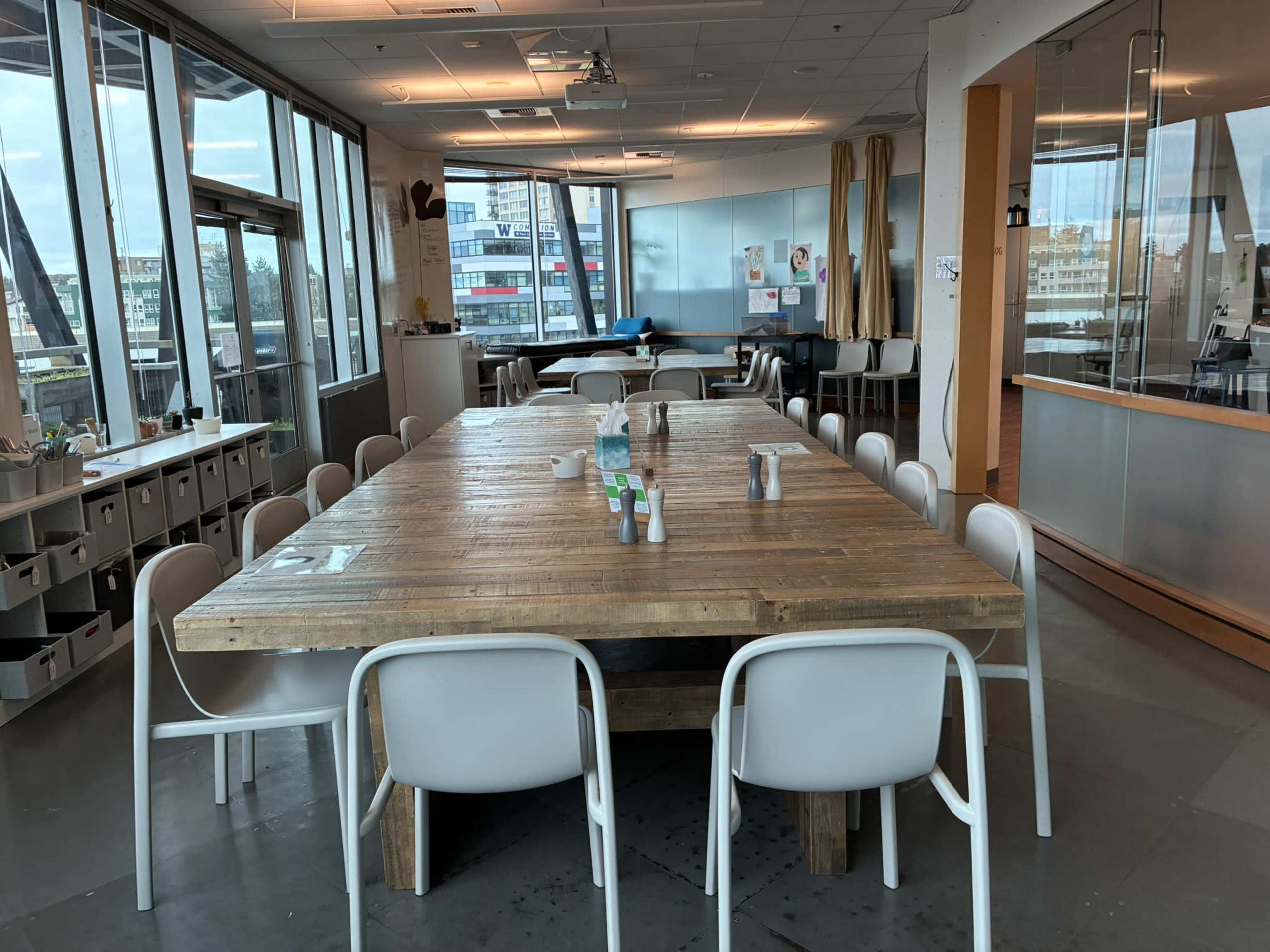 A large, wooden dining table surrounded by white chairs is set in a spacious, well-lit room with large windows.