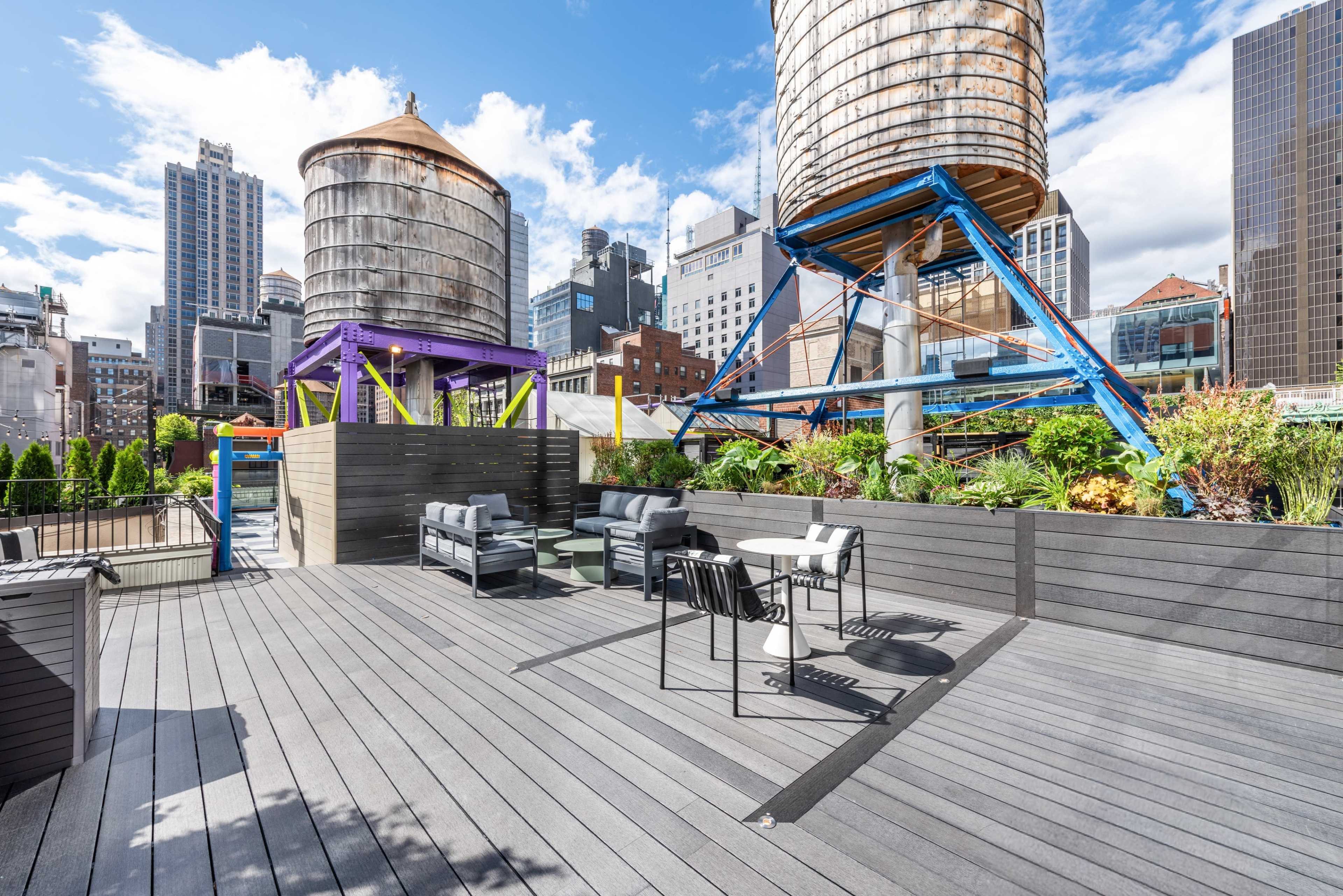The image shows a rooftop deck with modern furniture, surrounded by green plants and featuring two large water towers in an urban skyline.