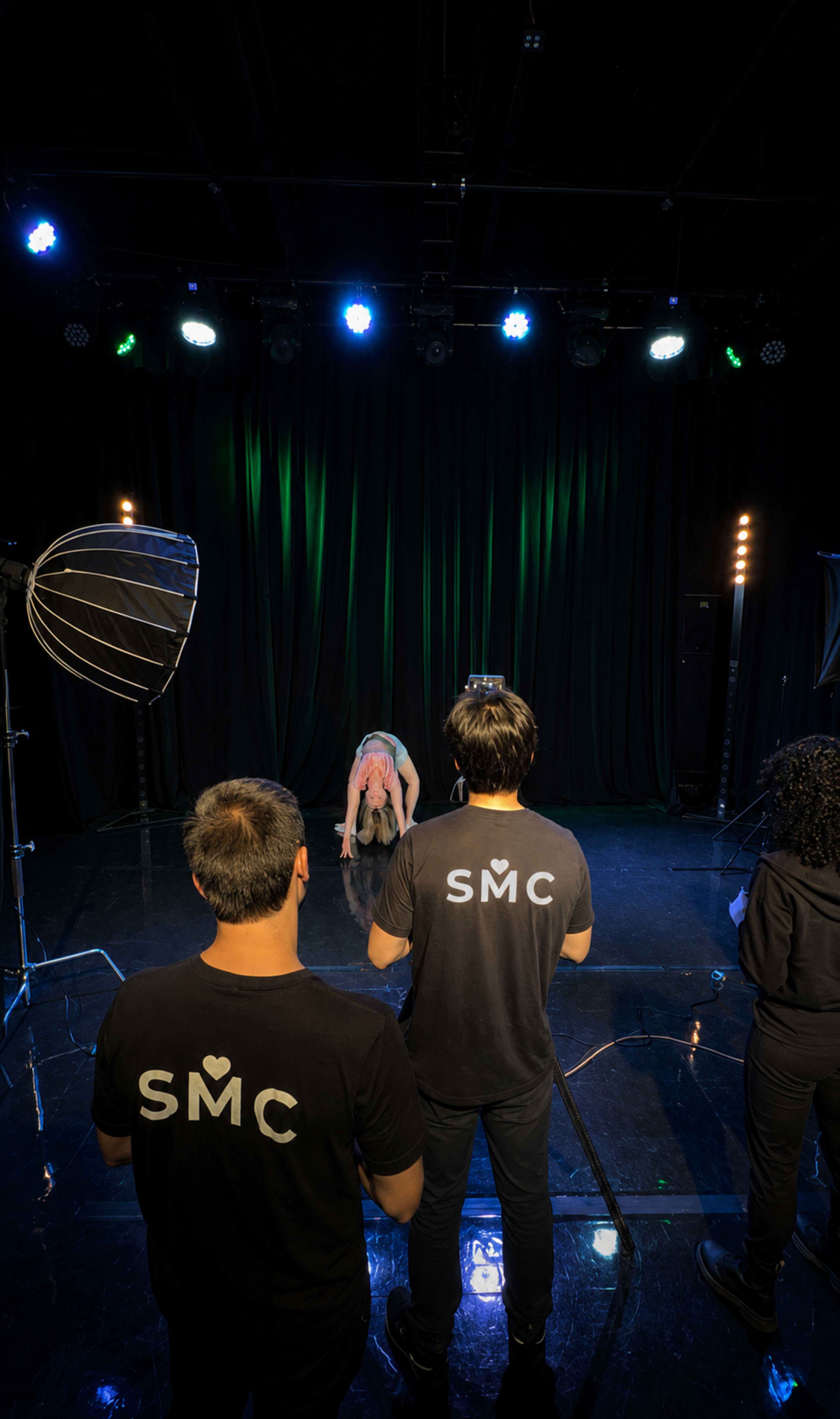 A dancer performs on stage while three crew members observe from behind, wearing matching black shirts with "SMC" printed on them.