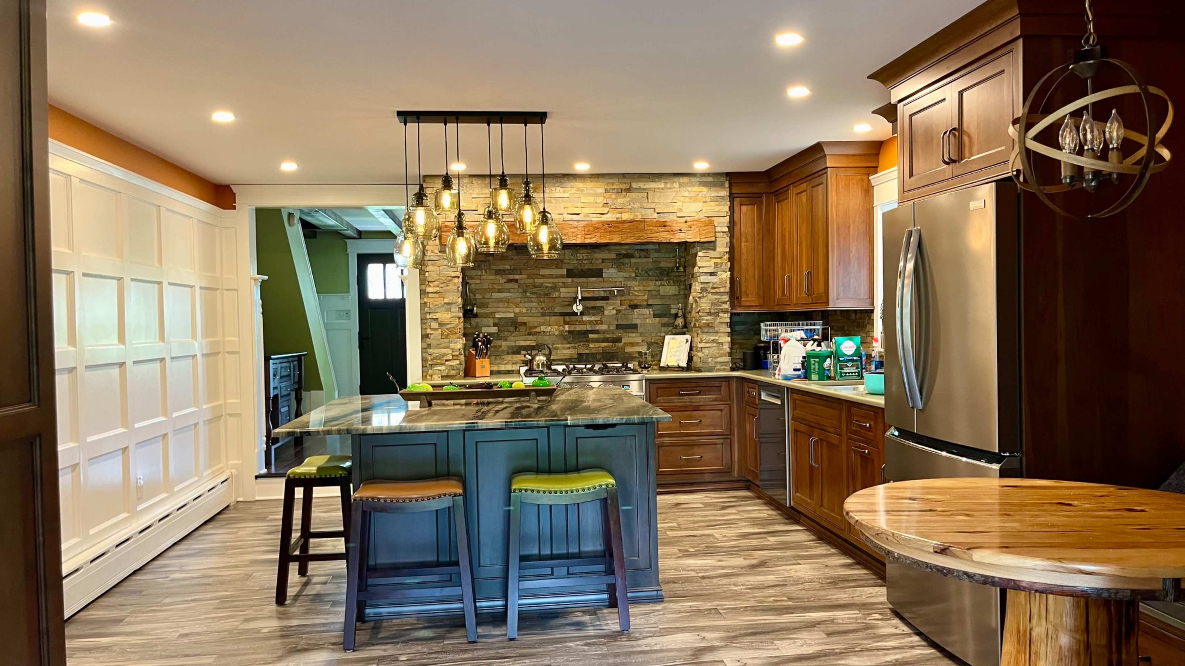 The image shows a modern kitchen with wooden cabinetry, a stone accent wall, an island with seating, and pendant lighting.