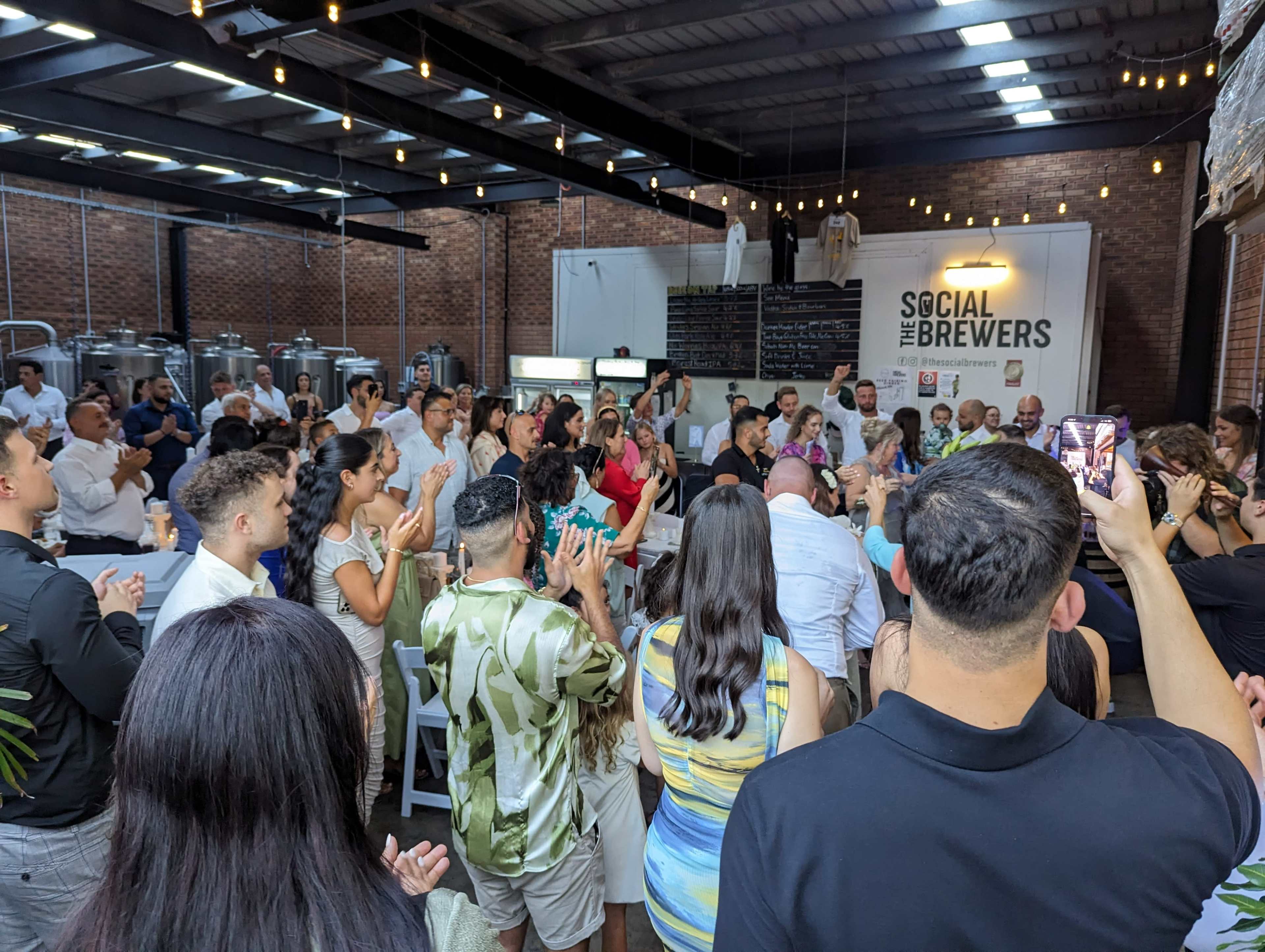 A large group of people is gathered in a brewery, clapping and facing a speaker at the front of the room.