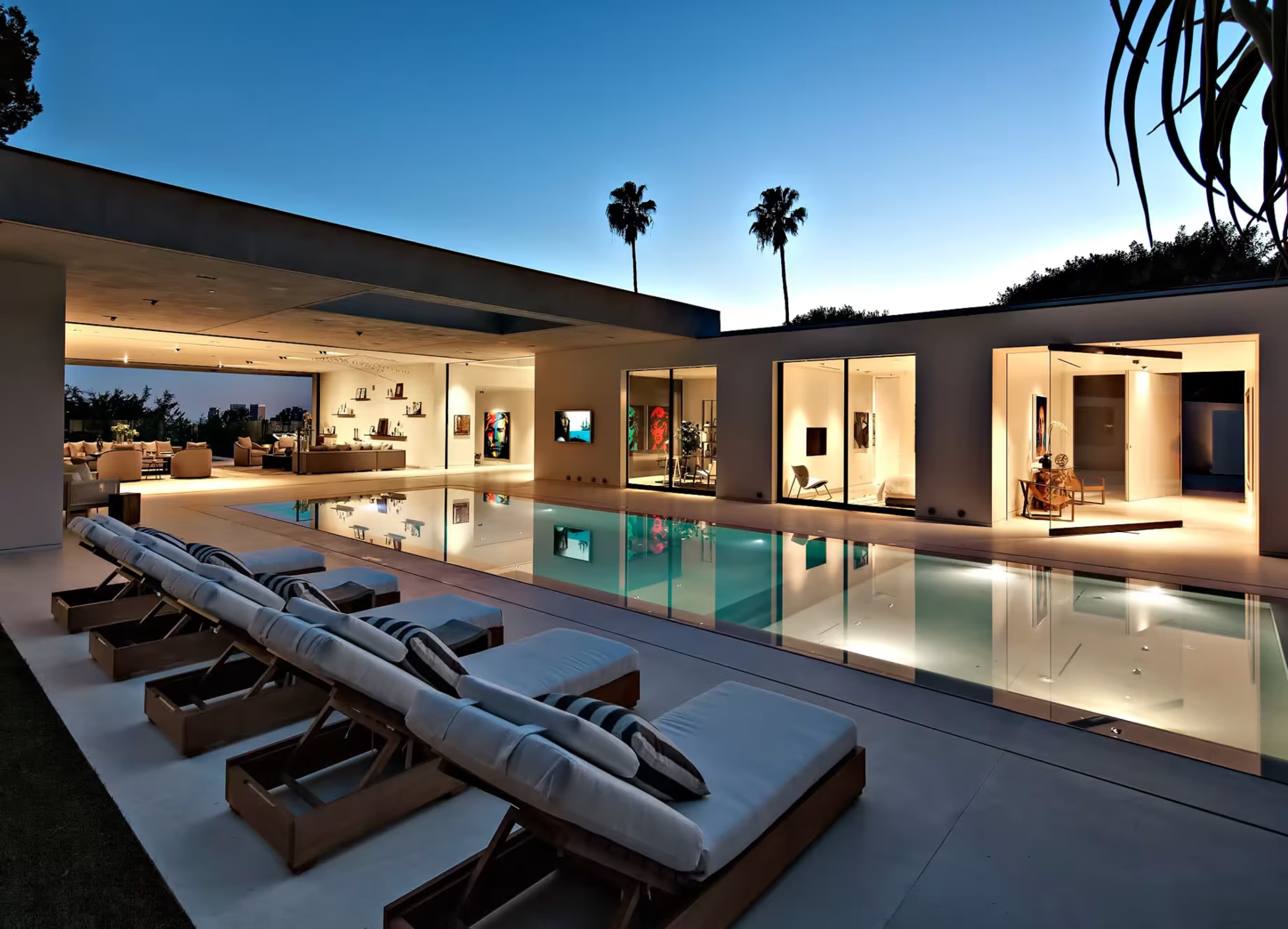 The image shows a modern house with large glass walls surrounding a swimming pool, featuring lounge chairs beside the water and palm trees silhouetted against a twilight sky.