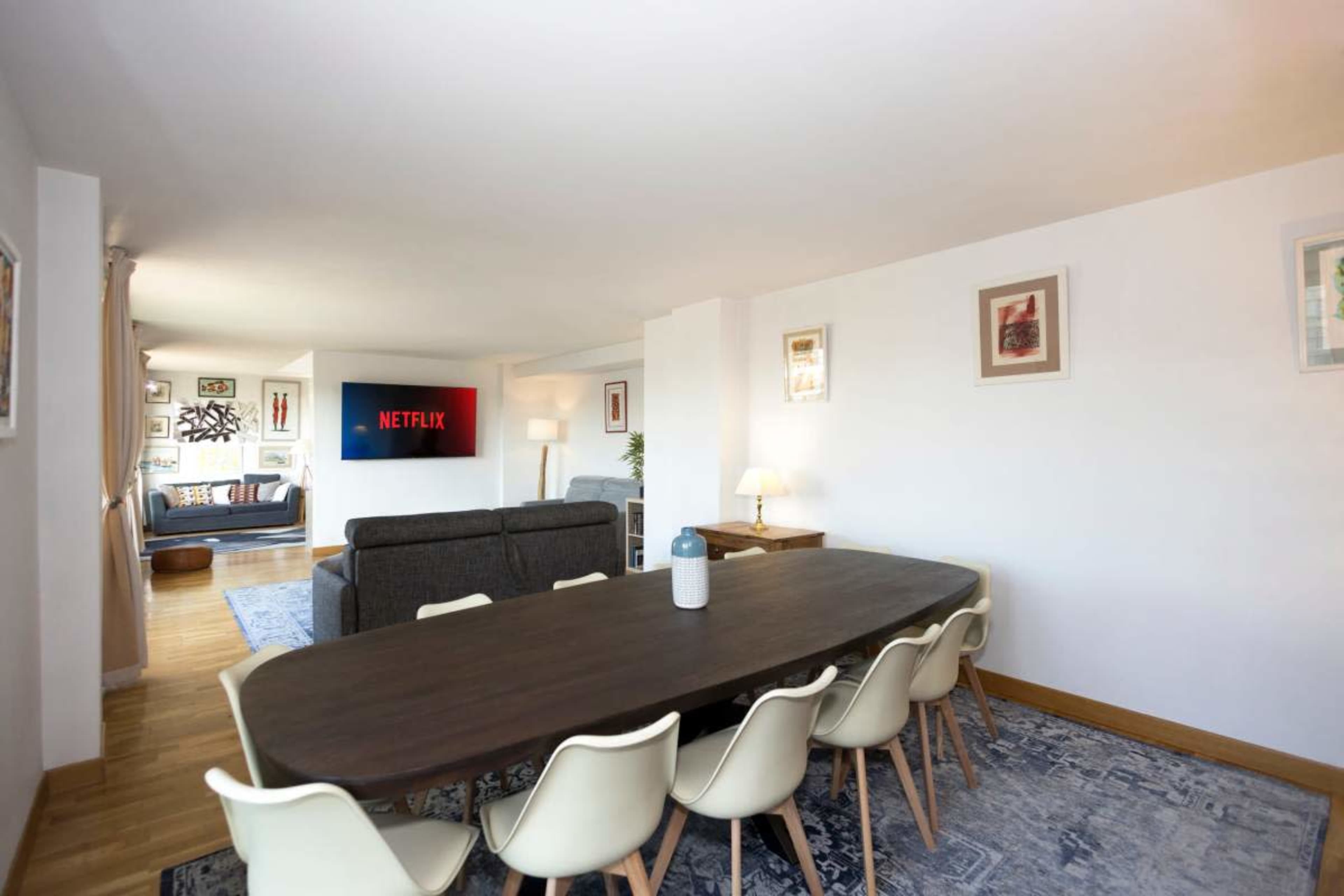 A modern living space features a dining table with eight chairs, a couch in the background, and a wall-mounted television displaying the Netflix logo.