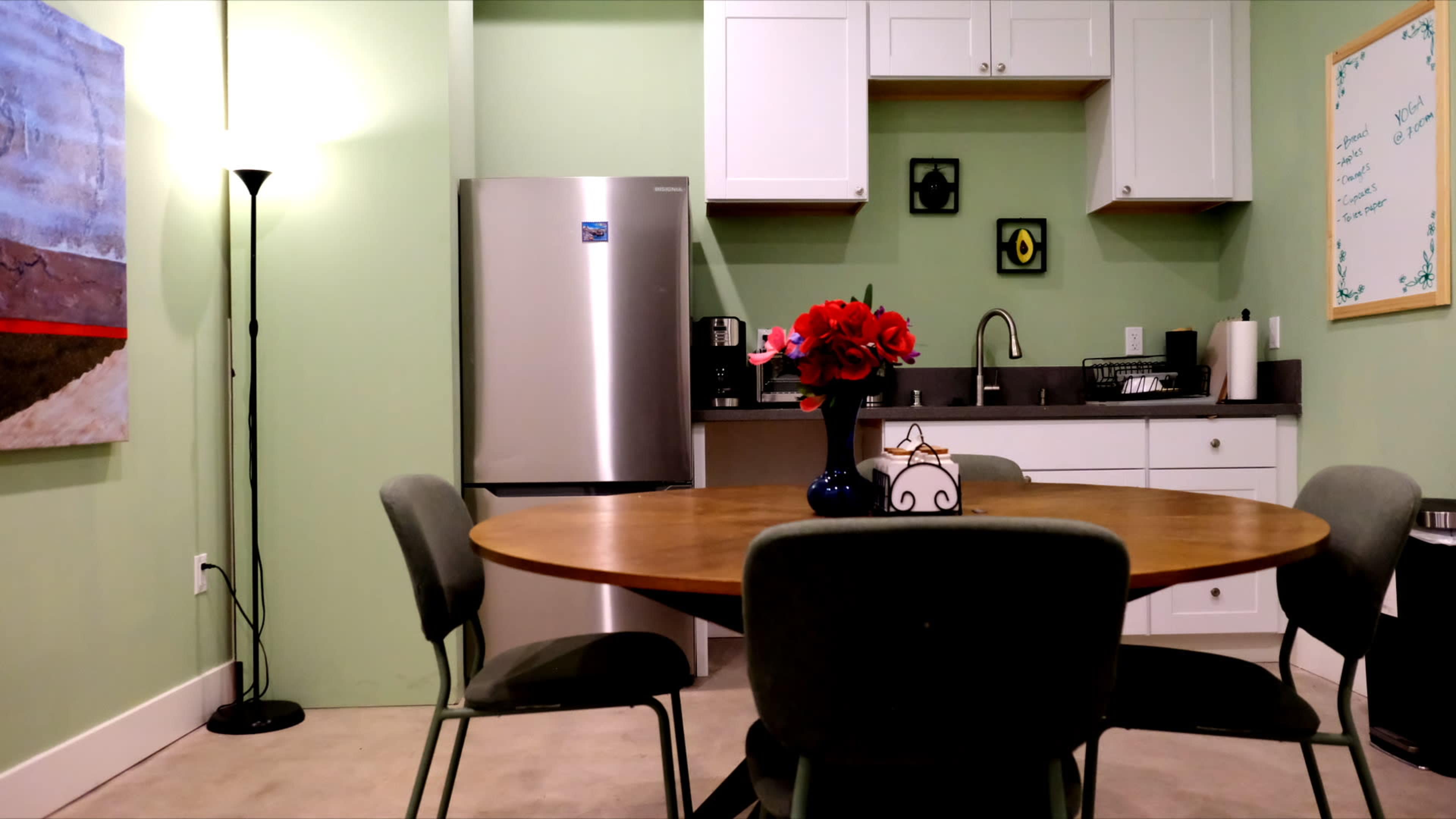 The image shows a kitchen area with a round wooden table surrounded by chairs, a refrigerator, a sink, and cabinets on the wall, with a vase of red flowers as the centerpiece.