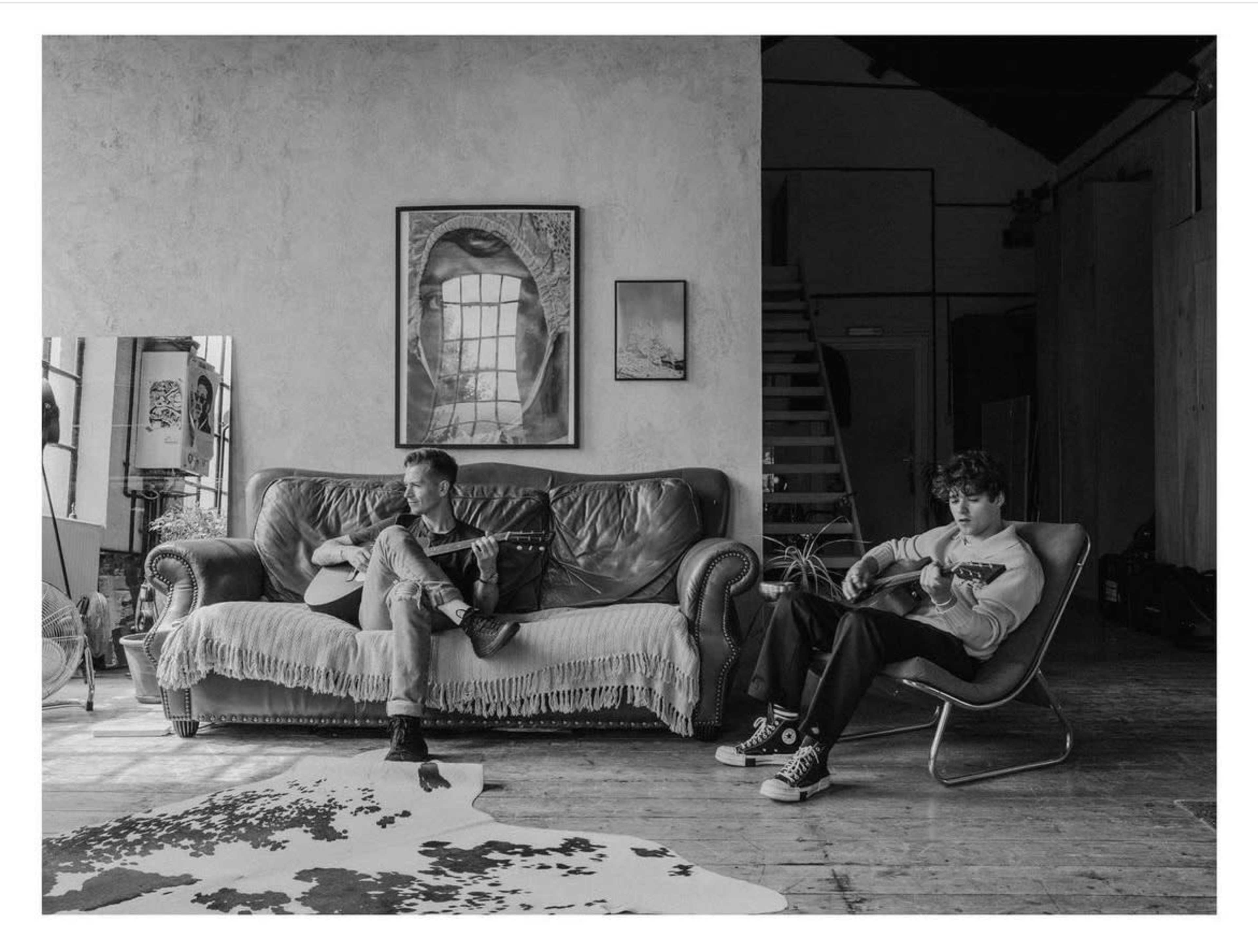 Two musicians sit in a spacious room, one on a sofa playing a guitar and the other in a chair strumming an acoustic guitar.