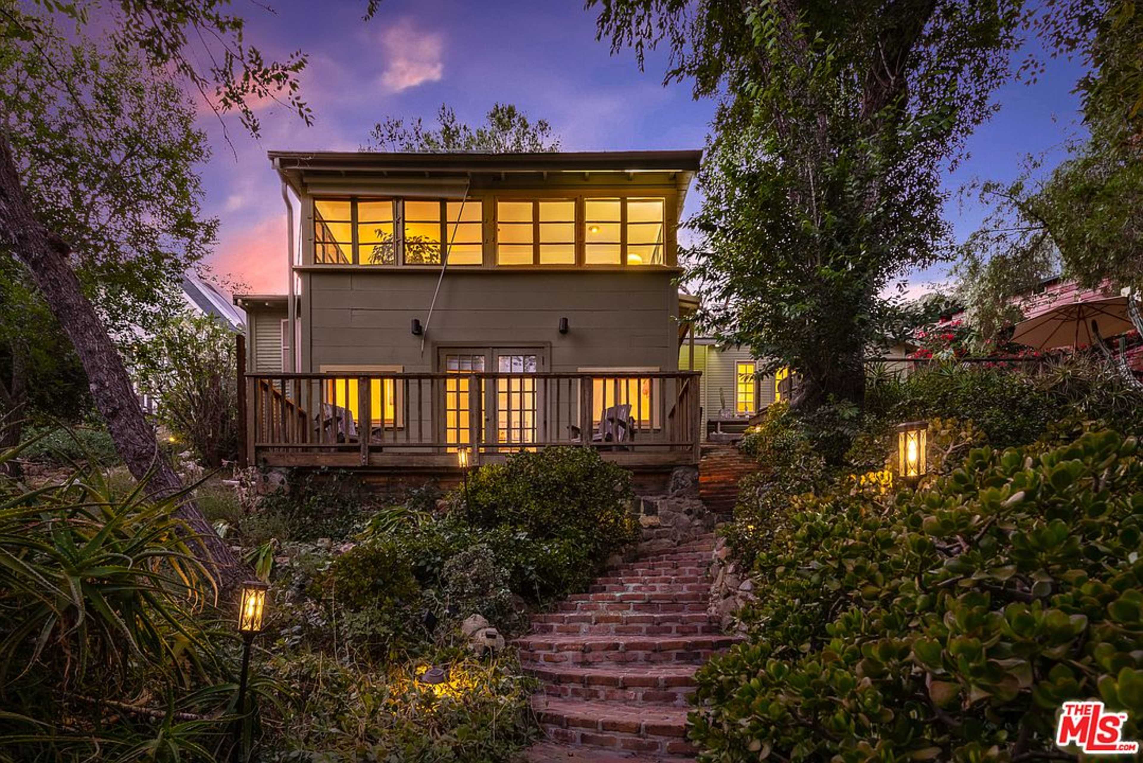 A two-story green house is surrounded by garden plants, with a brick pathway leading to a wooden deck illuminated by outdoor lights.
