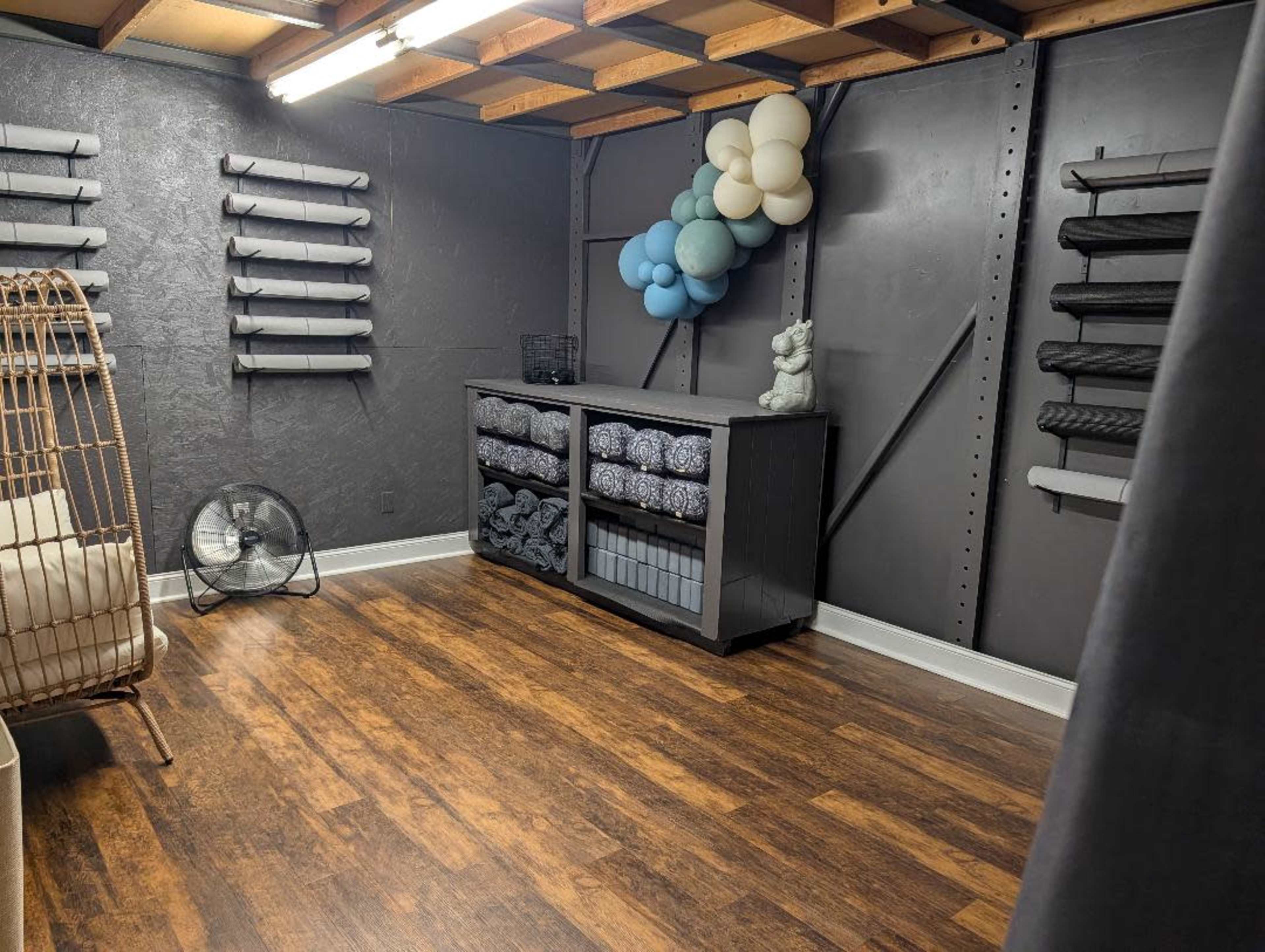 The image depicts a tidy room with a dark-colored wall, wooden flooring, a storage unit with neatly organized items, and balloons arranged in one corner.