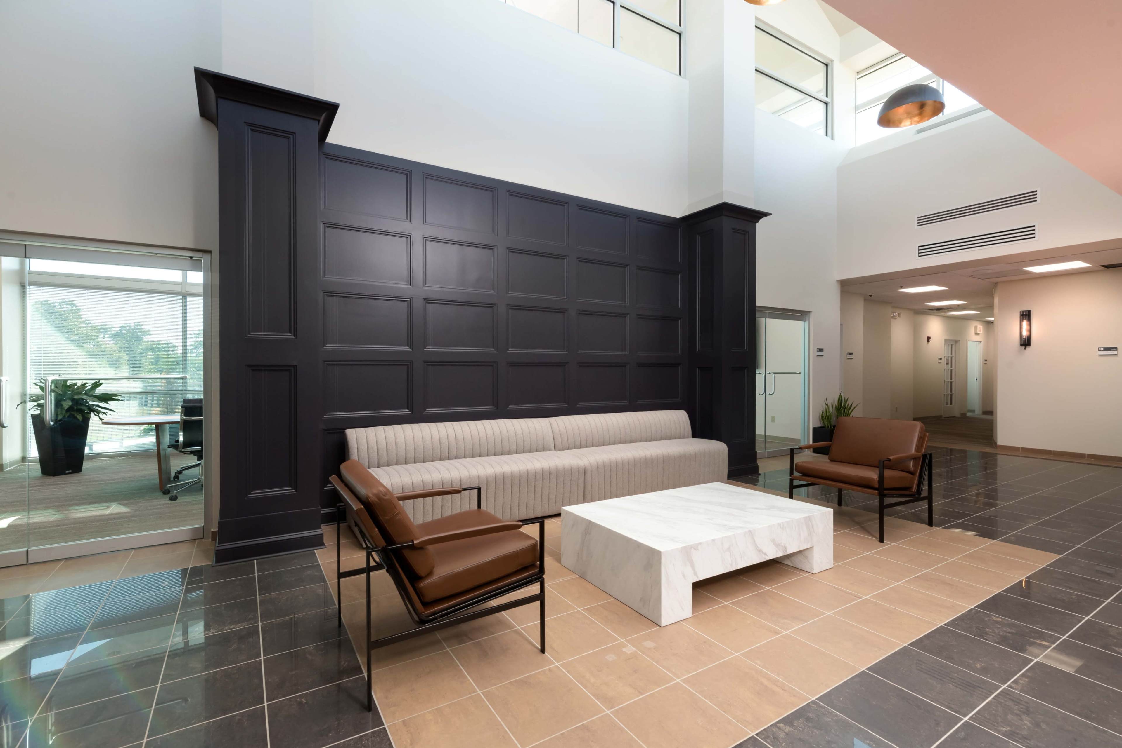 A modern lobby area featuring a gray sofa, two brown arm chairs, and a marble coffee table, all set against a dark paneled wall.