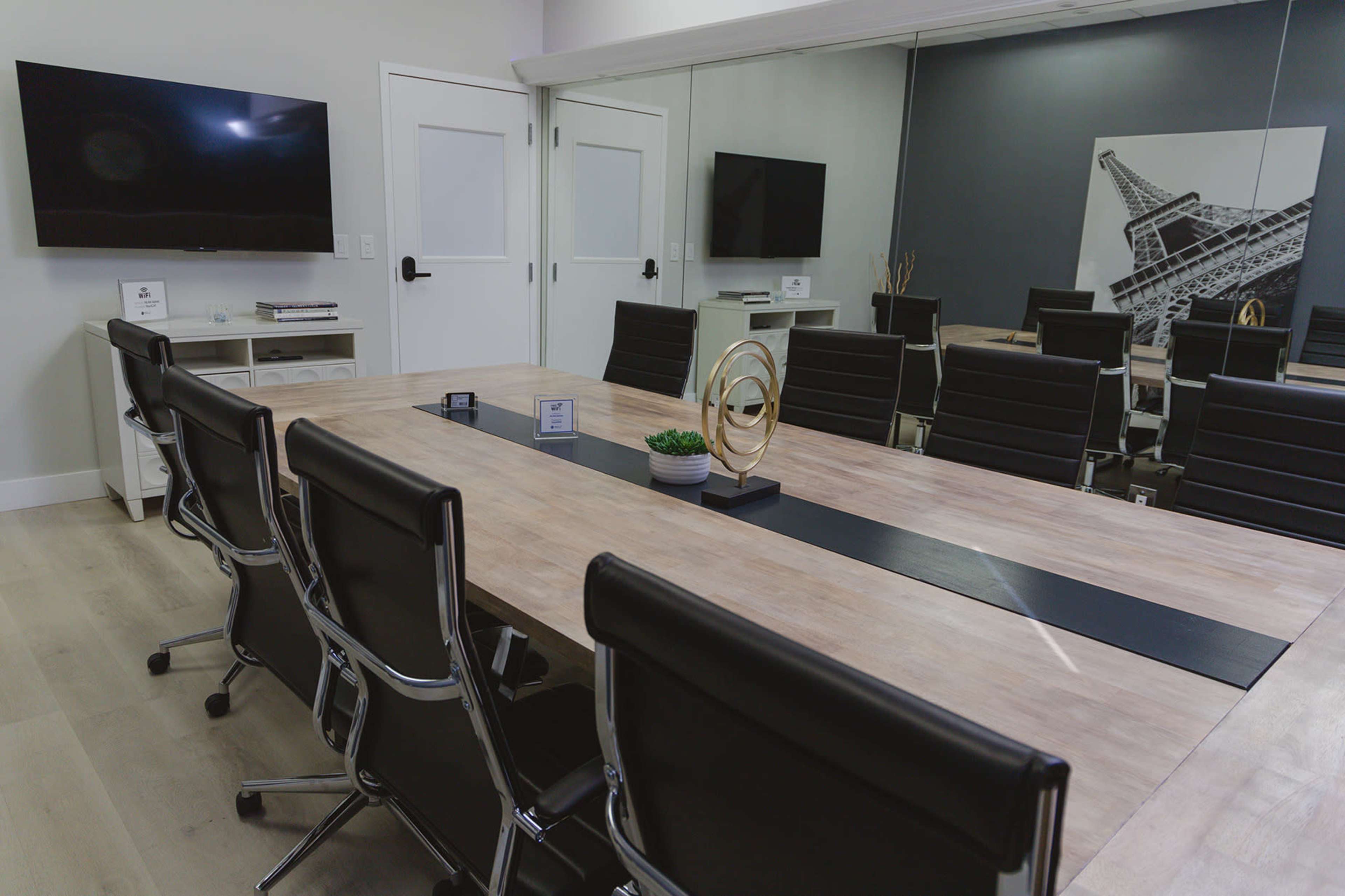 A modern conference room features a long wooden table surrounded by black leather chairs, with two televisions mounted on the walls.
