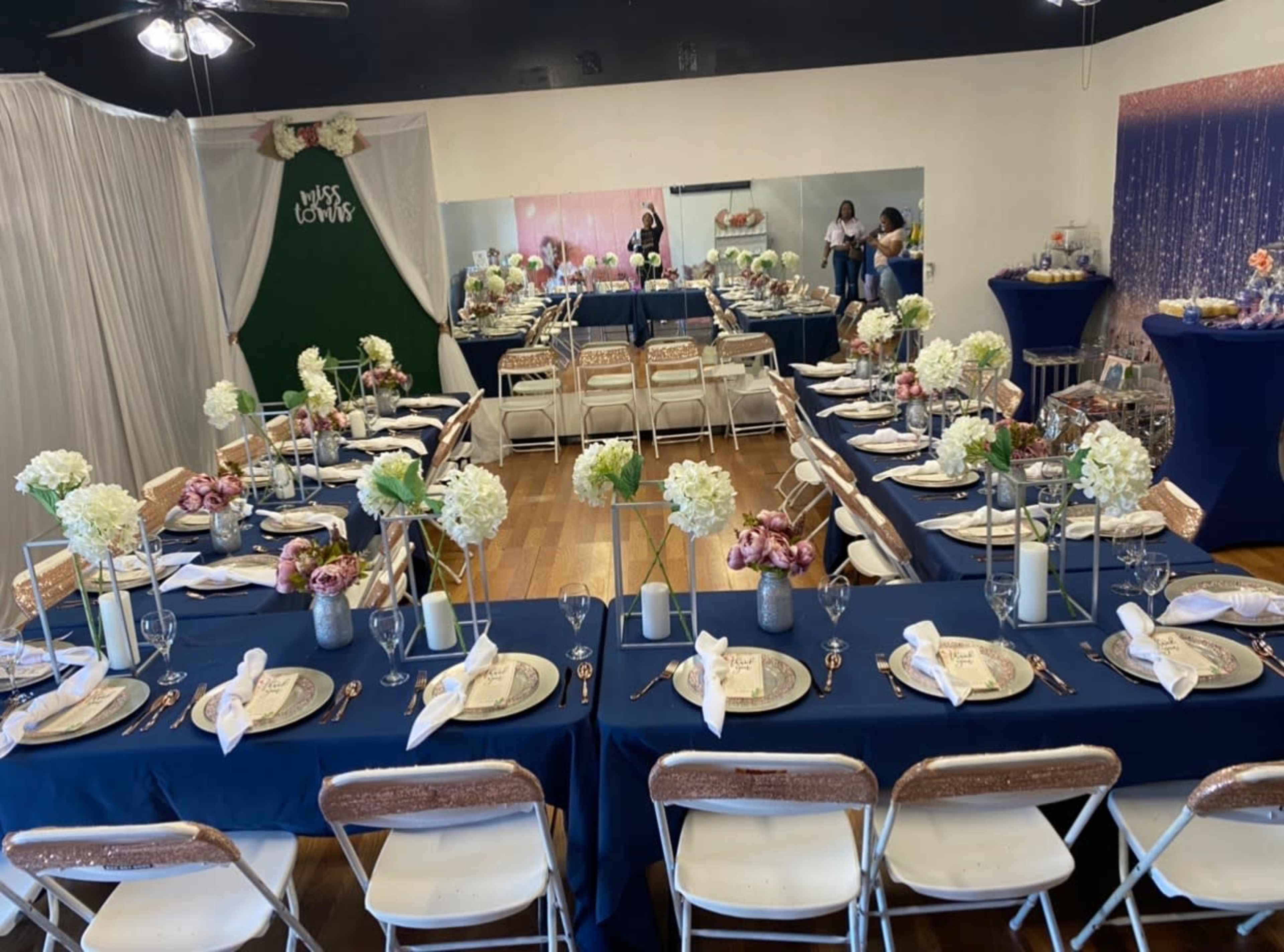 A banquet room is set up with long tables covered in blue tablecloths, arranged with floral centerpieces and place settings for a celebratory event.