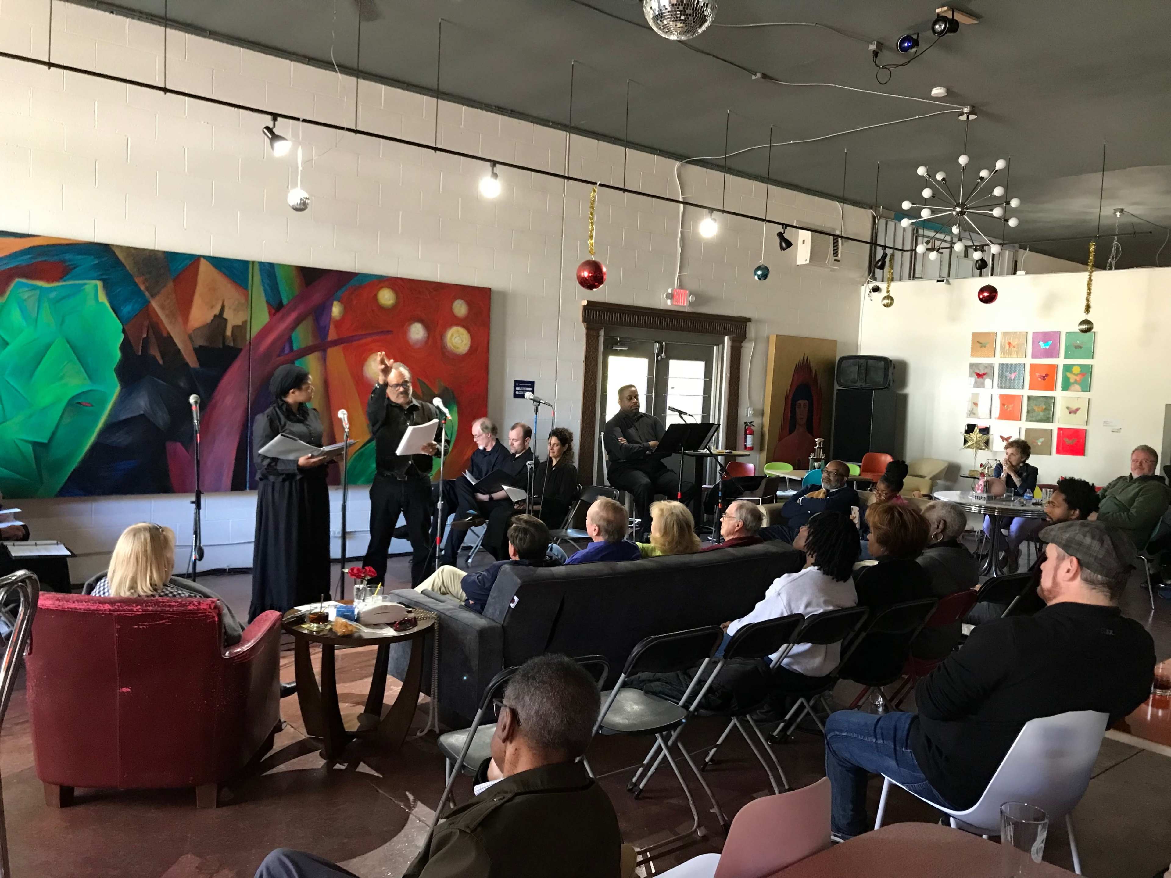 A group of people is gathered in a café, watching a performance taking place in front of colorful artwork on the wall.