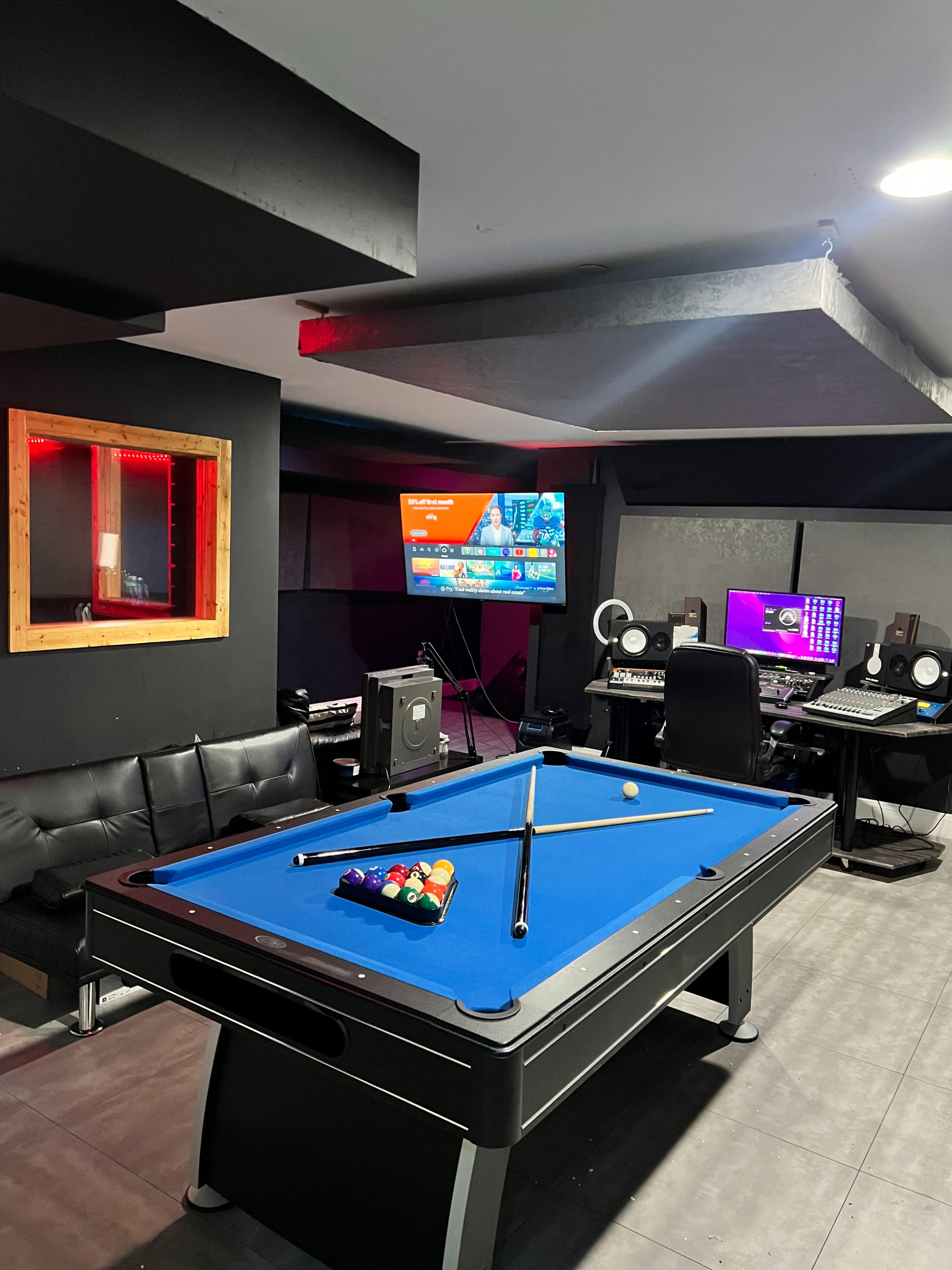The image shows a black billiards table with colorful balls arranged on it in a room featuring a seating area, a computer station with audio equipment, and a television mounted on the wall.